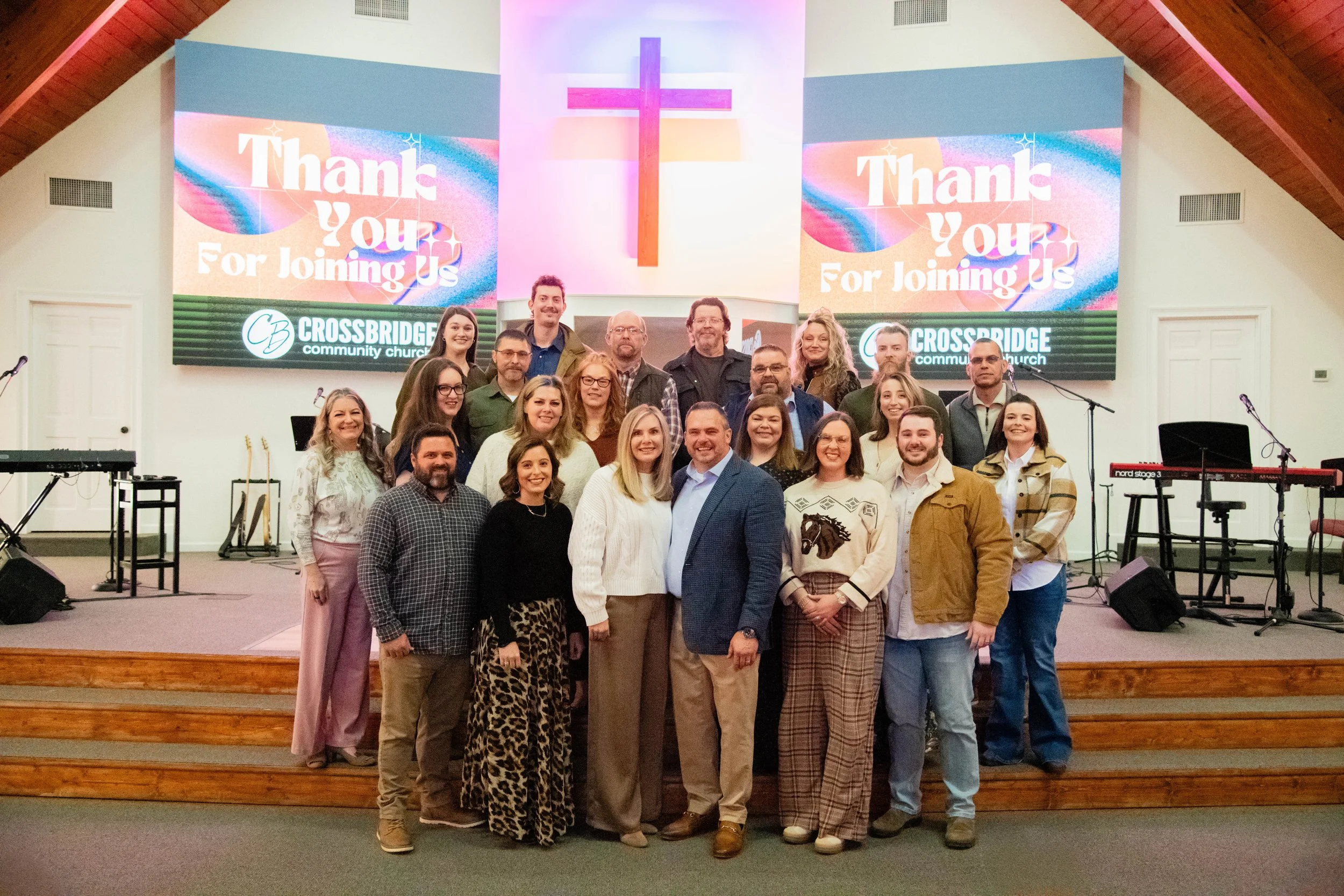 A group of people standing on a stage inside a church, with a cross on the wall behind them. The stage has musical equipment set up, including keyboards and microphones. Two large screens display a message of thanks from Crossbridge Community Church.