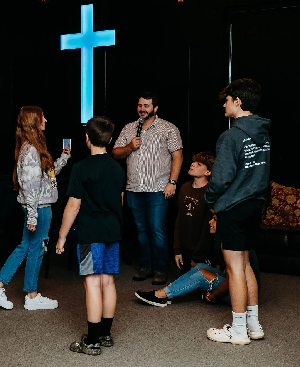 A group of young people and a man with a microphone inside a church with a lit cross on the wall. Some are standing, some are sitting on the floor. One girl is taking a photo or video with her phone.