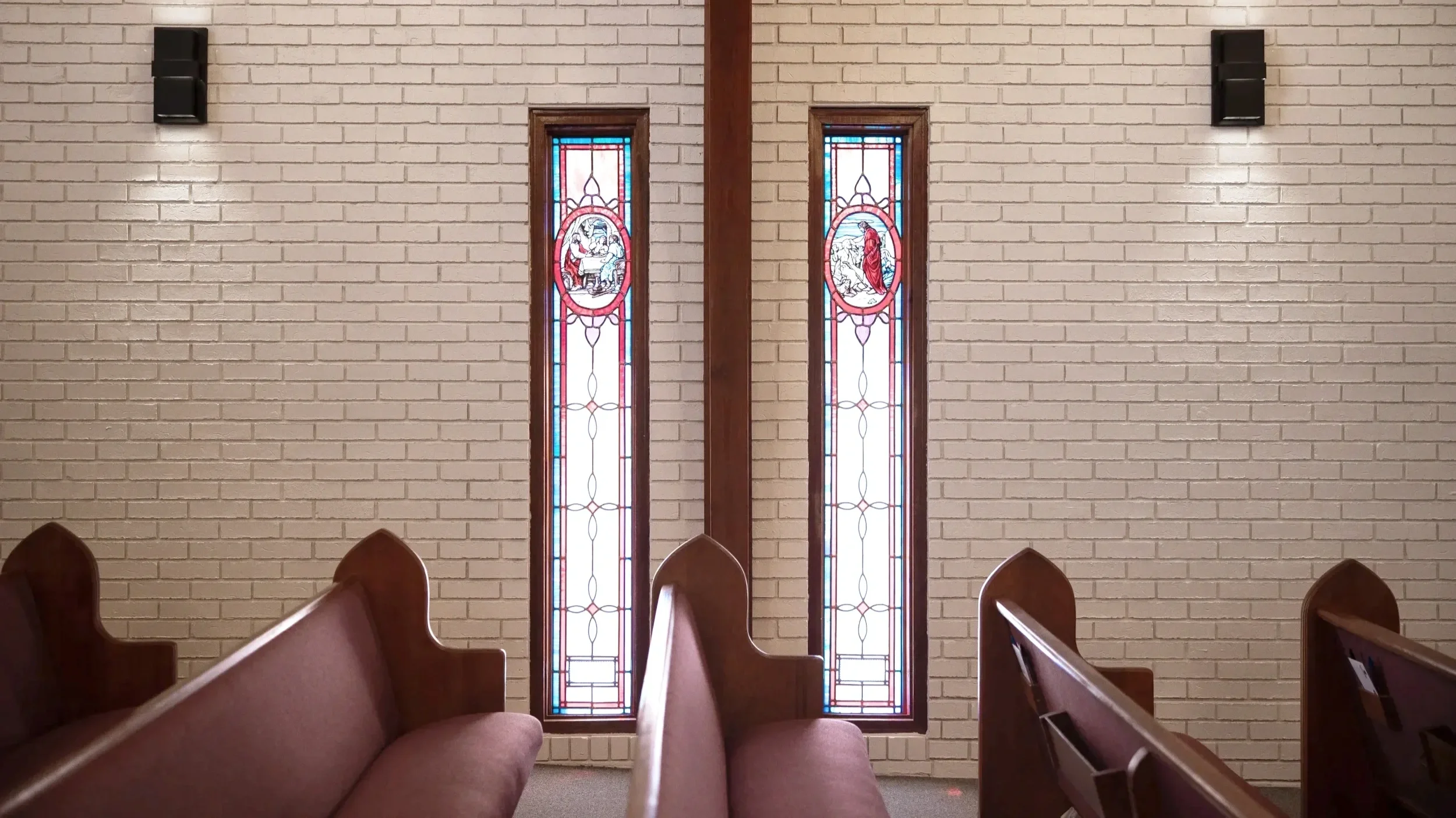 Inside a church with rows of wooden pews and two stained glass windows depicting religious scenes on a white brick wall.