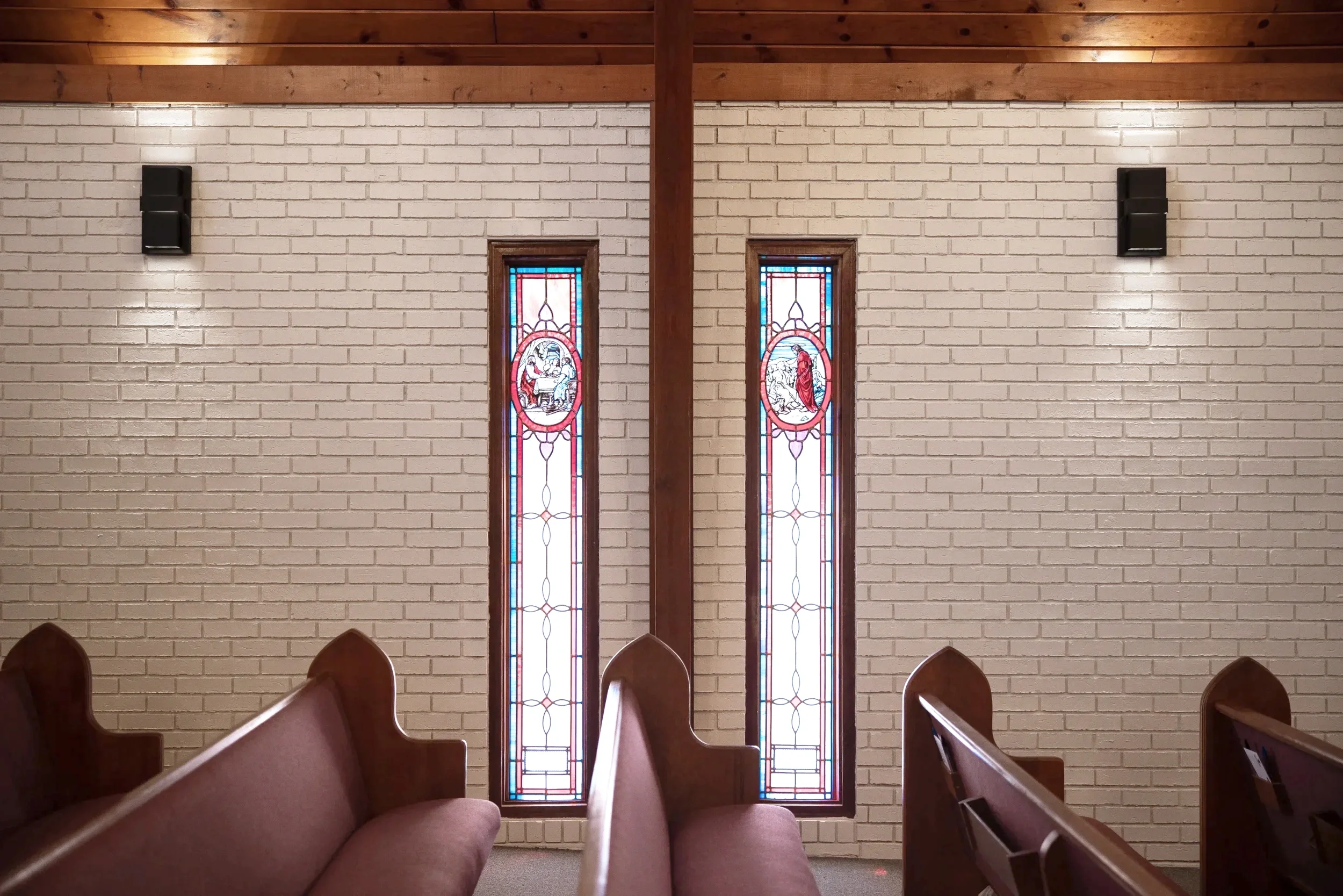 Interior of a church with beige brick walls, two tall stained glass windows with religious scenes, wooden pews, and wall-mounted speakers.
