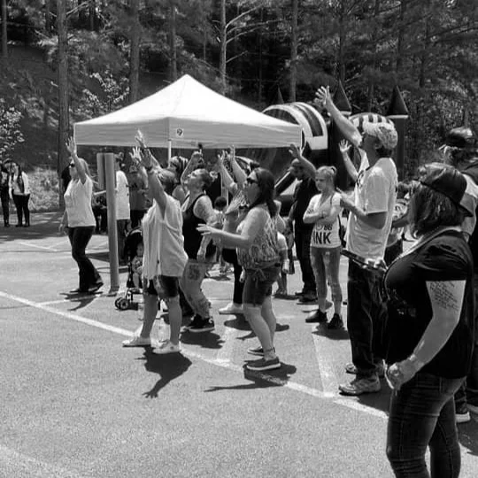 Group of people dancing and celebrating outdoors under a tent at a park or fair.