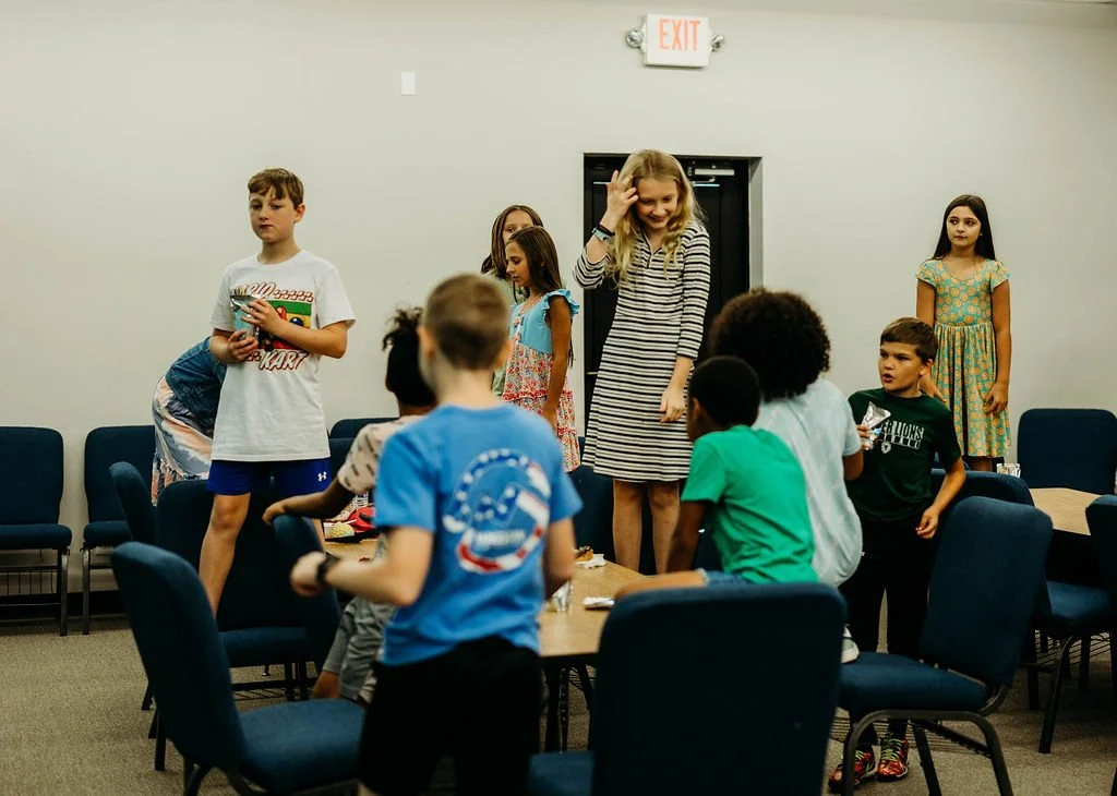 Children standing and sitting in a room with chairs and a table, some looking at each other, and others looking in different directions, with an exit sign above a doorway in the background.