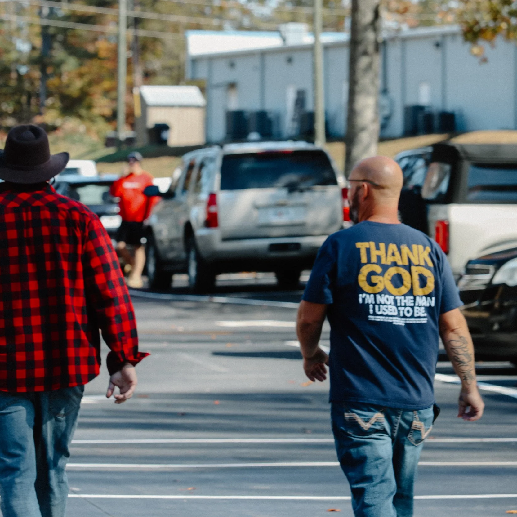 Two men walking in a parking lot with cars, one wearing a blue shirt with religious text and the other in a red and black checkered shirt and a hat, while a third person stands near the cars in the background.