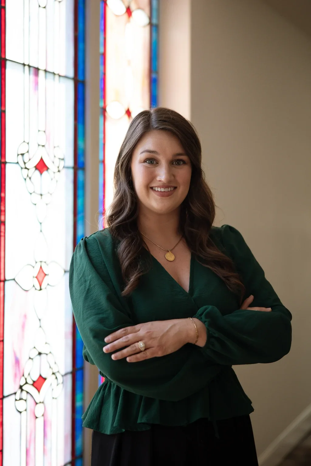 A young woman standing in front of stained glass windows inside a church or chapel. She has long, wavy brown hair, is smiling, and is wearing a dark green blouse with puffed sleeves and a gold necklace.