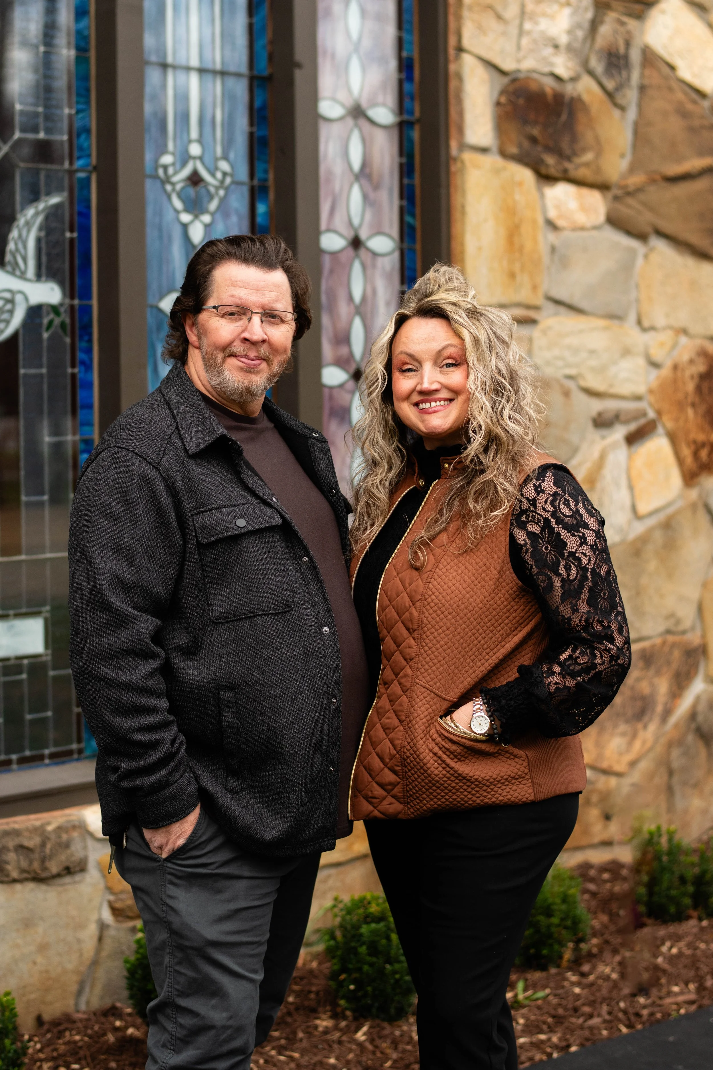 A man and woman standing outside in front of a stone wall and stained glass window, smiling at the camera.