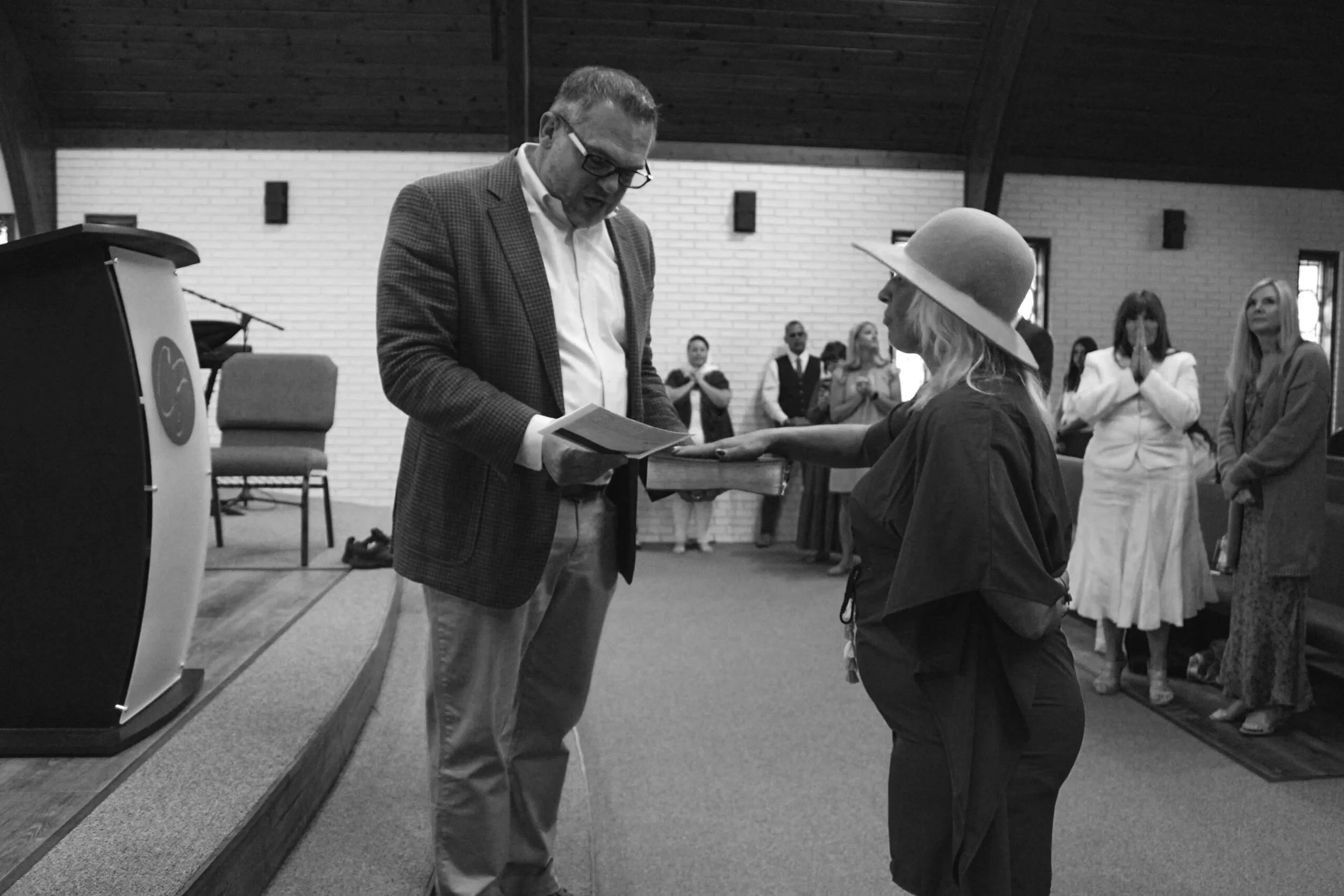 A woman in a black outfit and wide-brimmed hat receiving an award or certificate from a man in a blazer, with several people standing and praying in the background in a church setting.