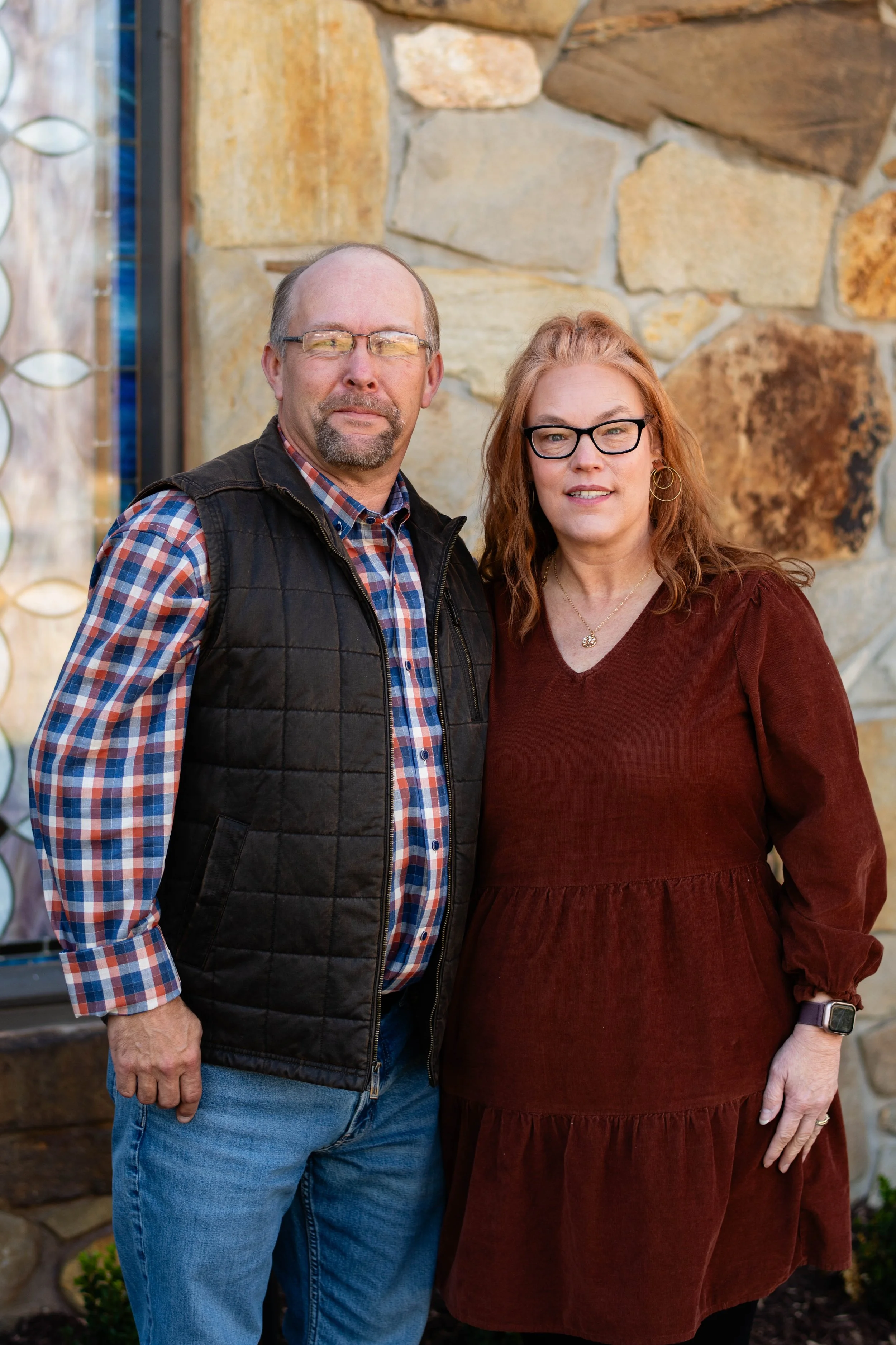 A man and woman standing outdoors in front of a stone wall. The man is wearing glasses, a plaid shirt, a black vest, and jeans. The woman has red hair, glasses, gold hoop earrings, and is dressed in a maroon dress. They are both facing the camera with neutral expressions.