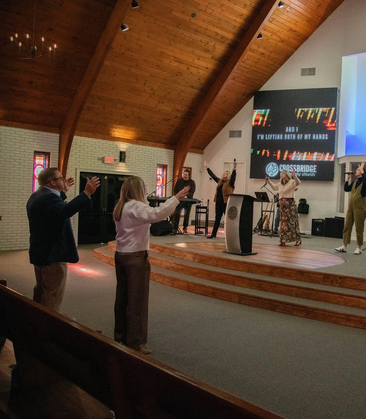 People worshiping in a church, with some standing and raising their hands, while others are singing or playing instruments on stage, inside a church with wooden ceiling and stained glass windows.
