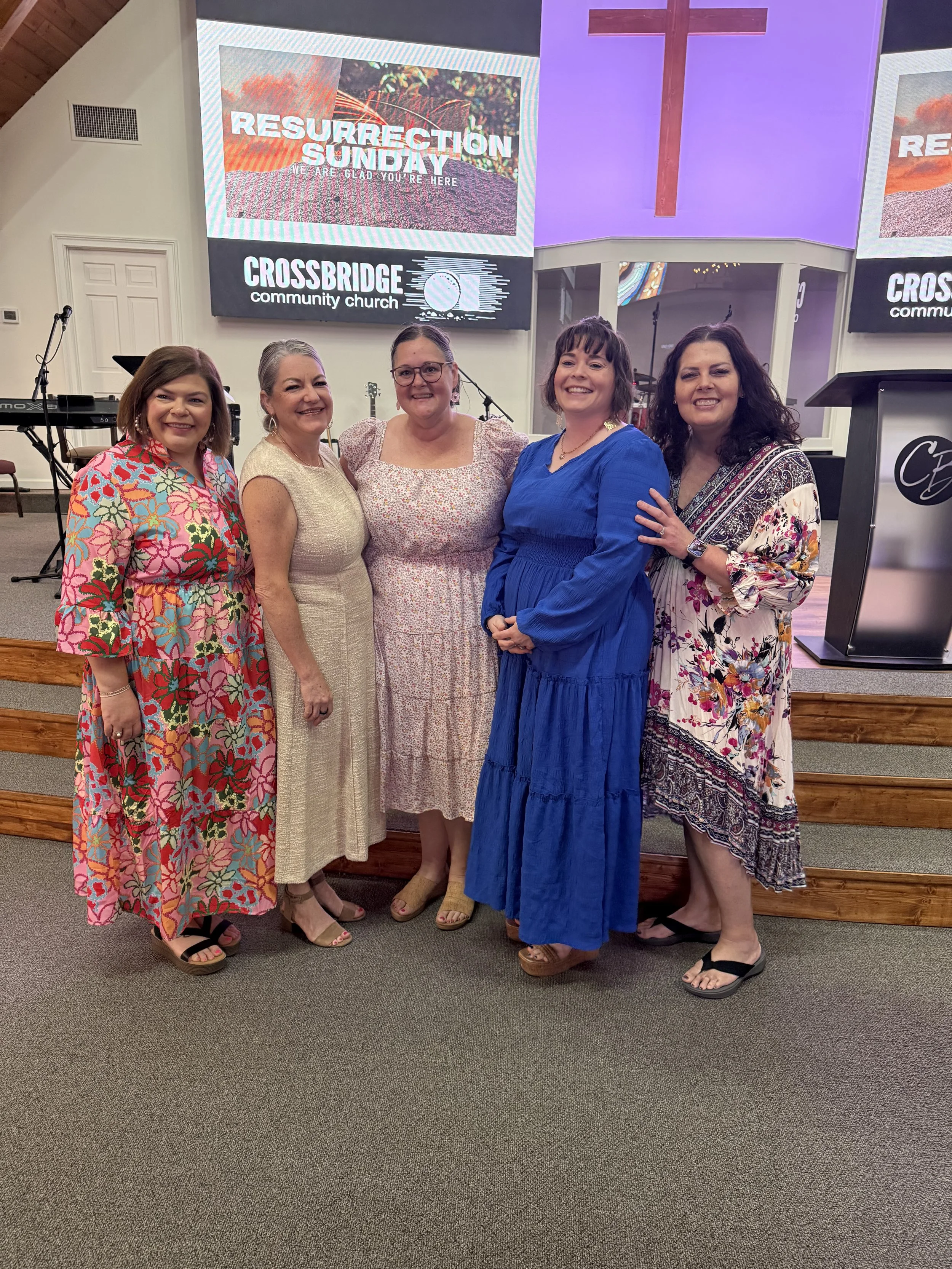 Five women standing together inside a church, smiling, with a large wooden cross and church screens displaying text about Resurrection Sunday in the background.