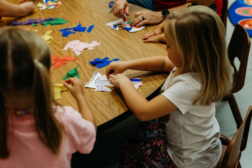 Children and adults sitting at a wooden table engaging in a craft activity with multicolored paper shapes.