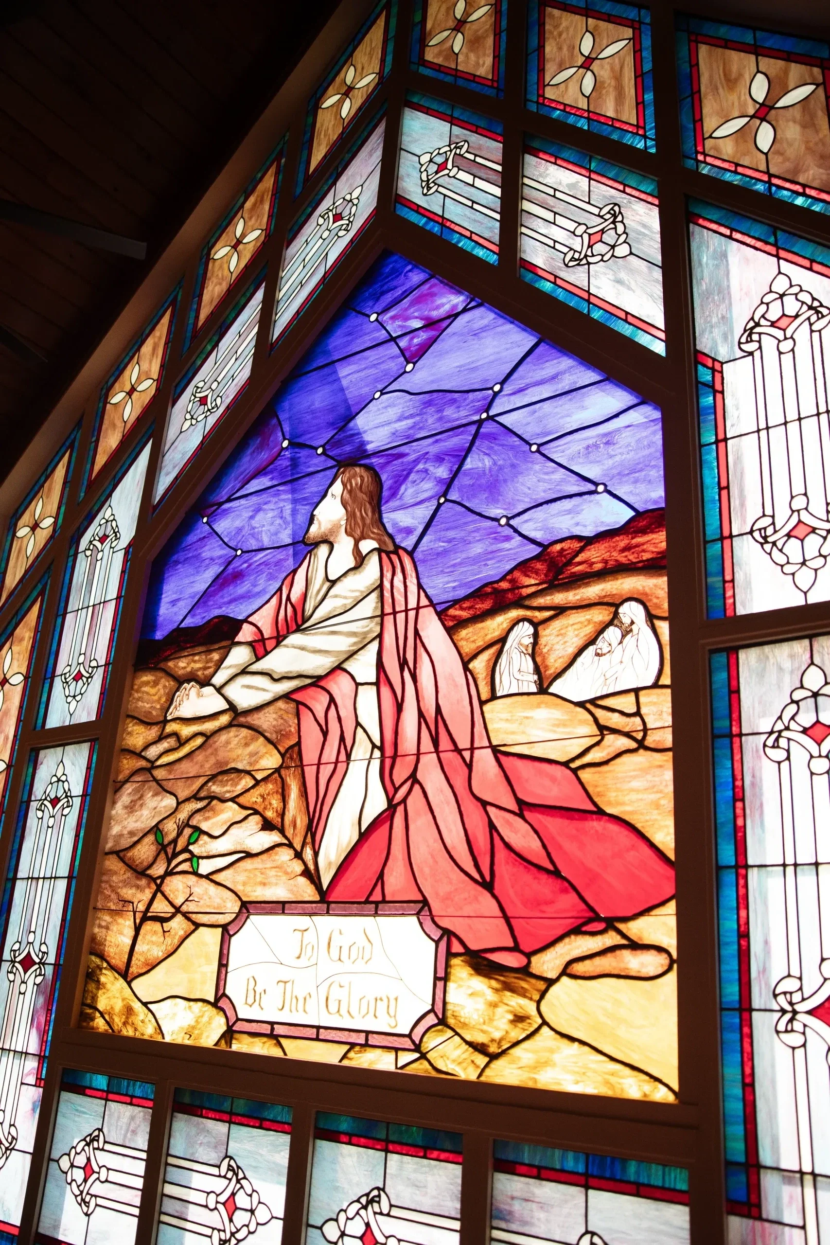 Stained glass window depicting Jesus Christ with a purple sky and mountains behind, with a sign reading 'To God Be The Glory' in front.