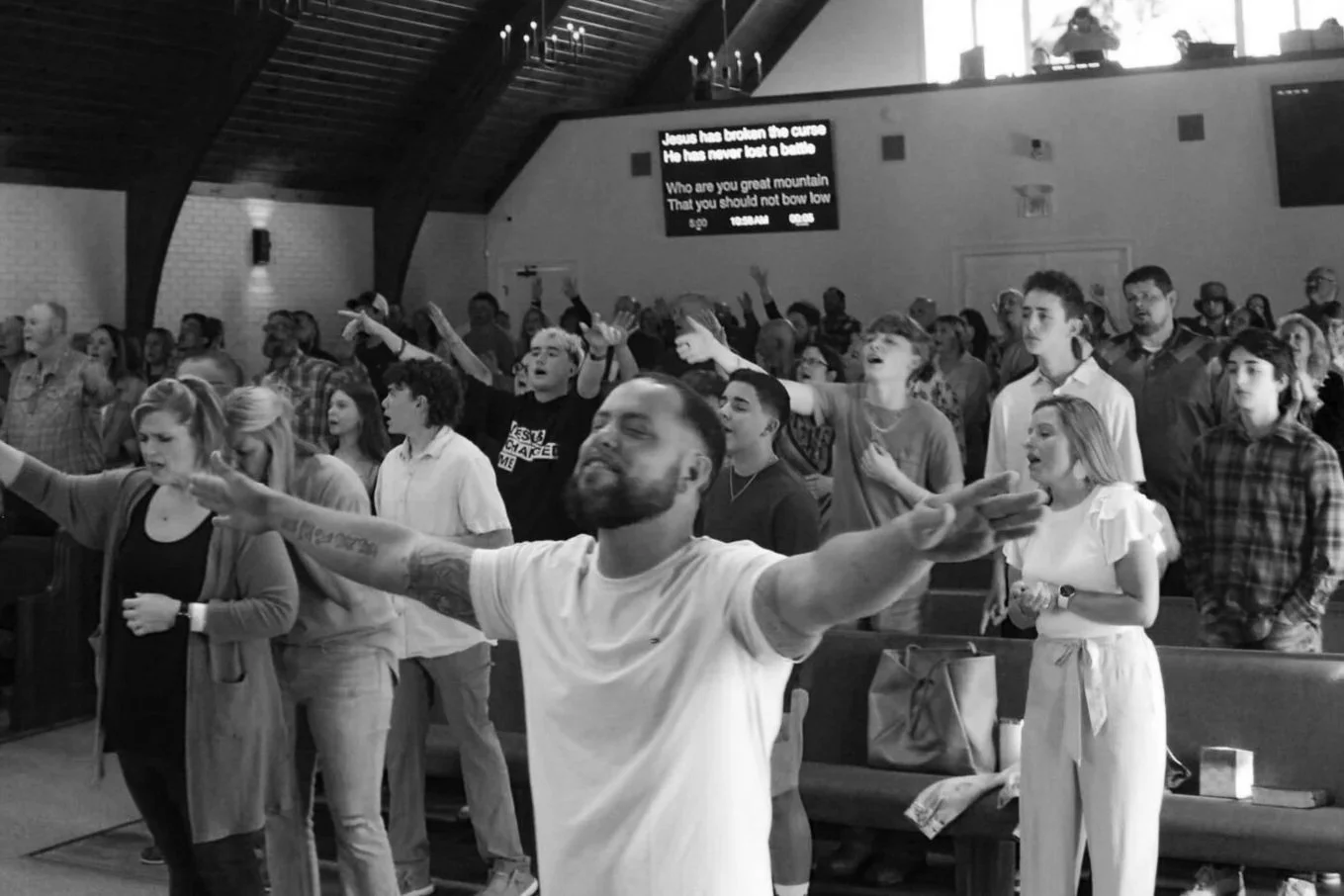A diverse group of people standing with arms raised and eyes closed in a church congregation, with a man in the foreground with arms outstretched and a beard, and a digital screen in the background displaying lyrics.