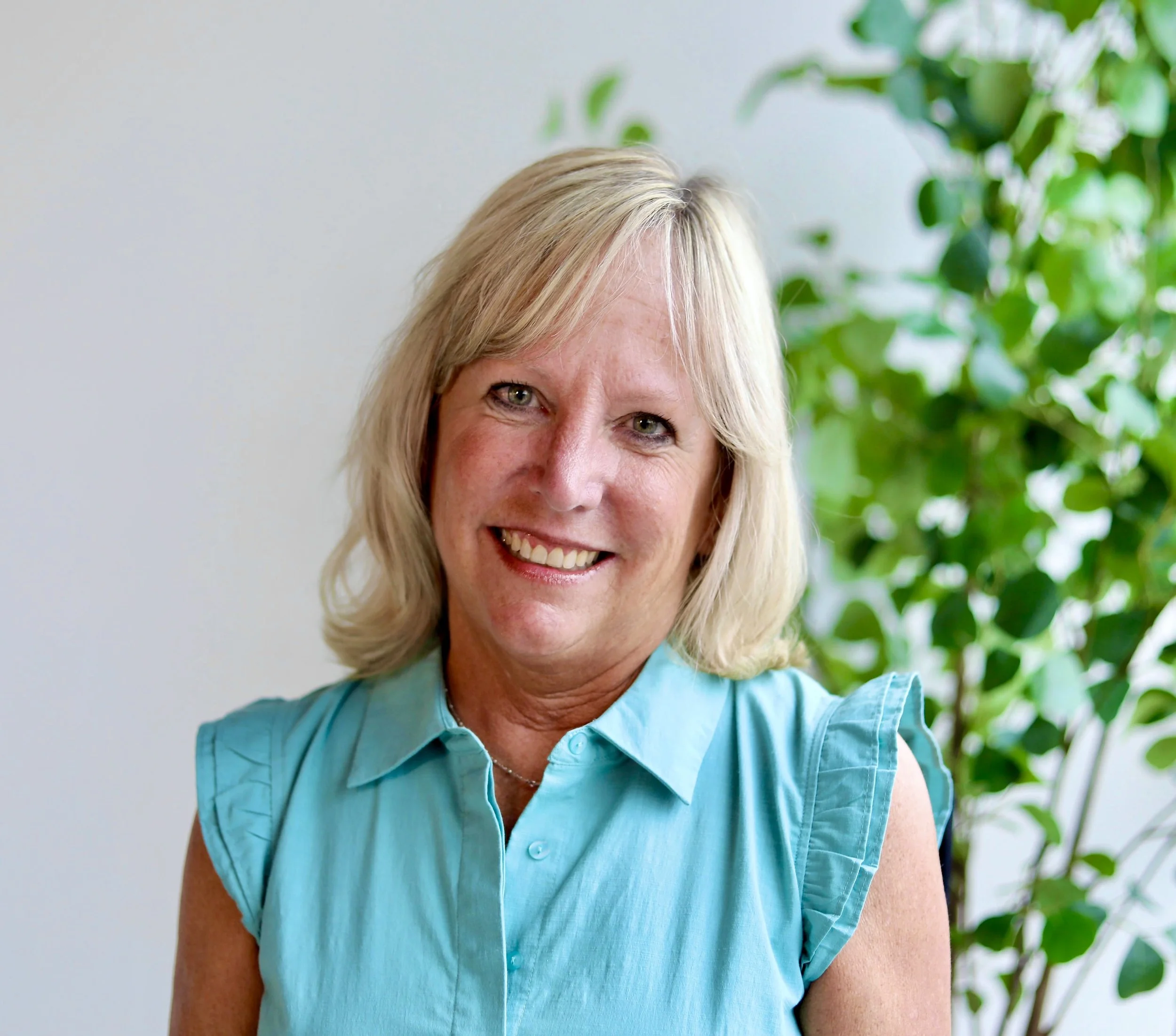 Tammy Rice, M.Ed., wearing a light blue sleeveless blouse, smiling indoors with green foliage in the background.