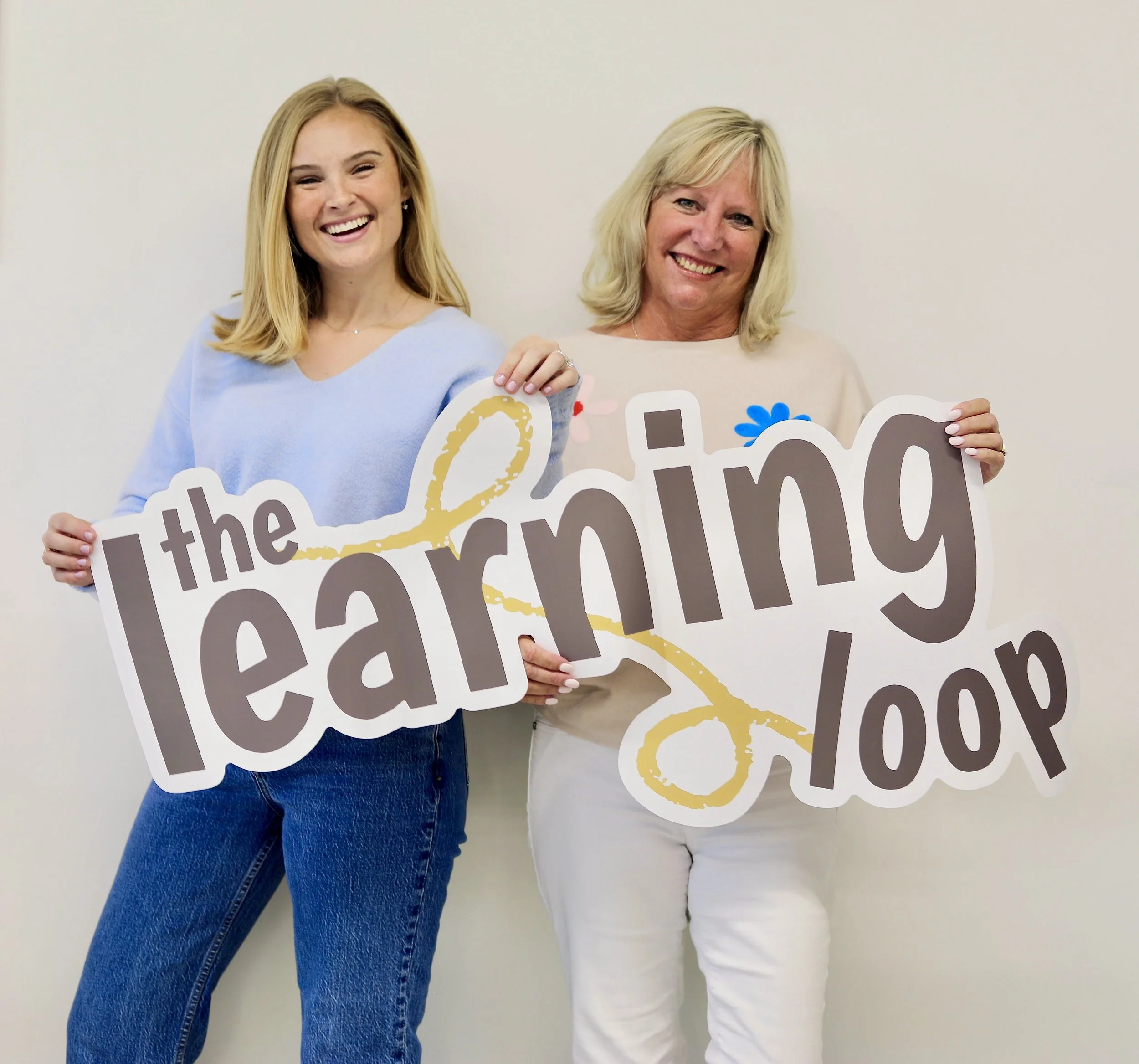 Shannon and Tammy smiling and holding a sign that reads 'The Learning Loop' in front of a plain white wall.