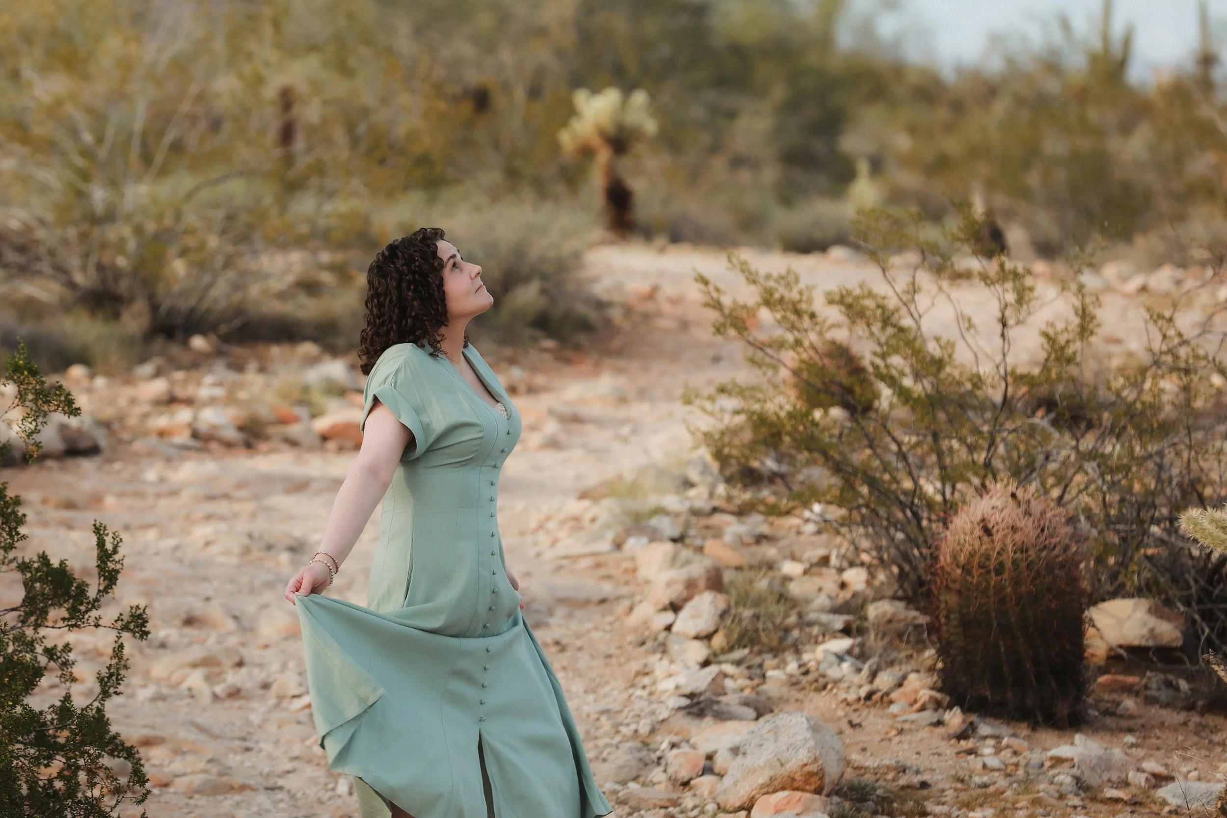 A woman with curly hair in a vintage light green dress standing outdoors in a desert landscape with sparse shrubs and rocks, looking upward.
