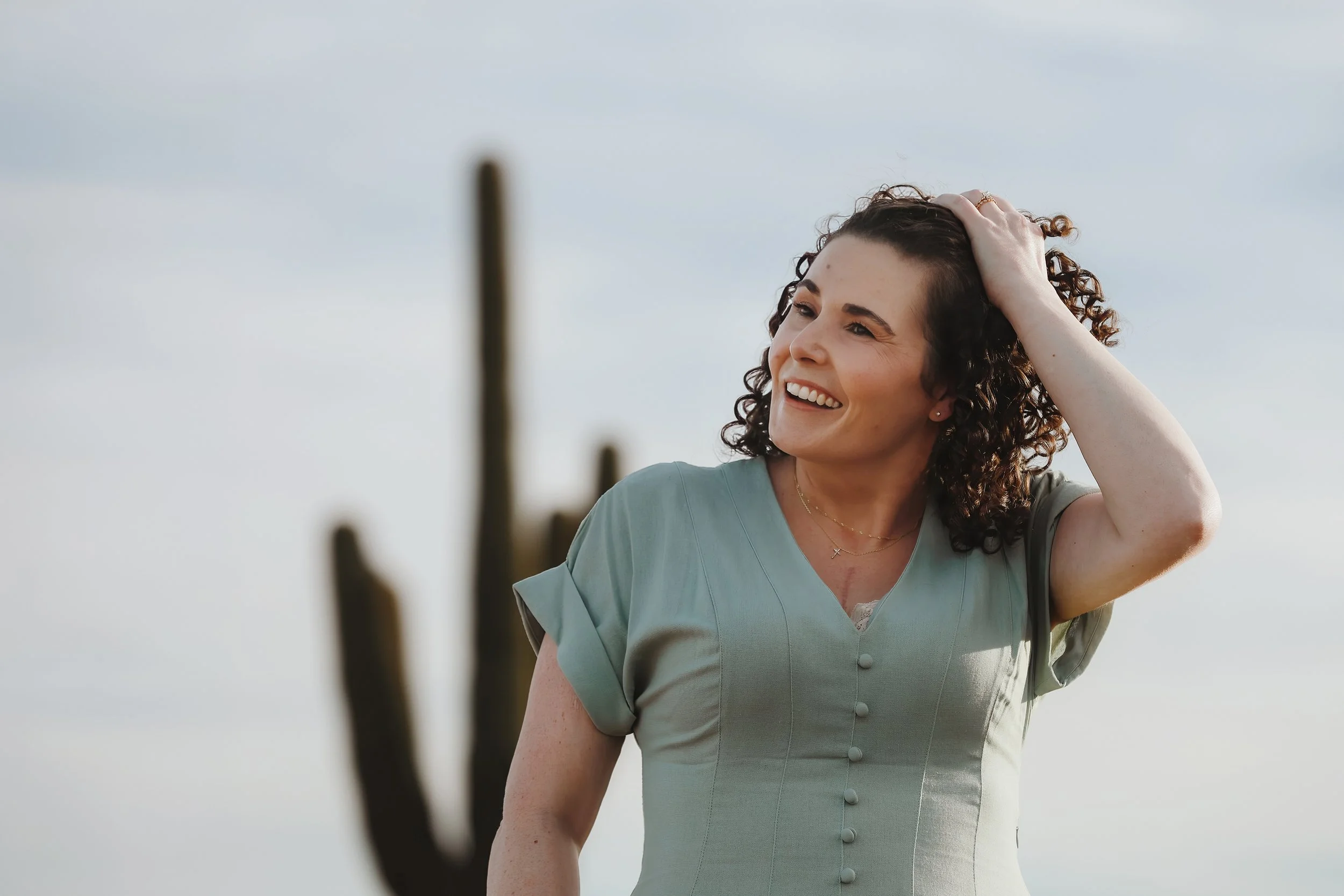 A woman with curly hair smiling and touching her head, standing outdoors with a cloudy sky in the background.