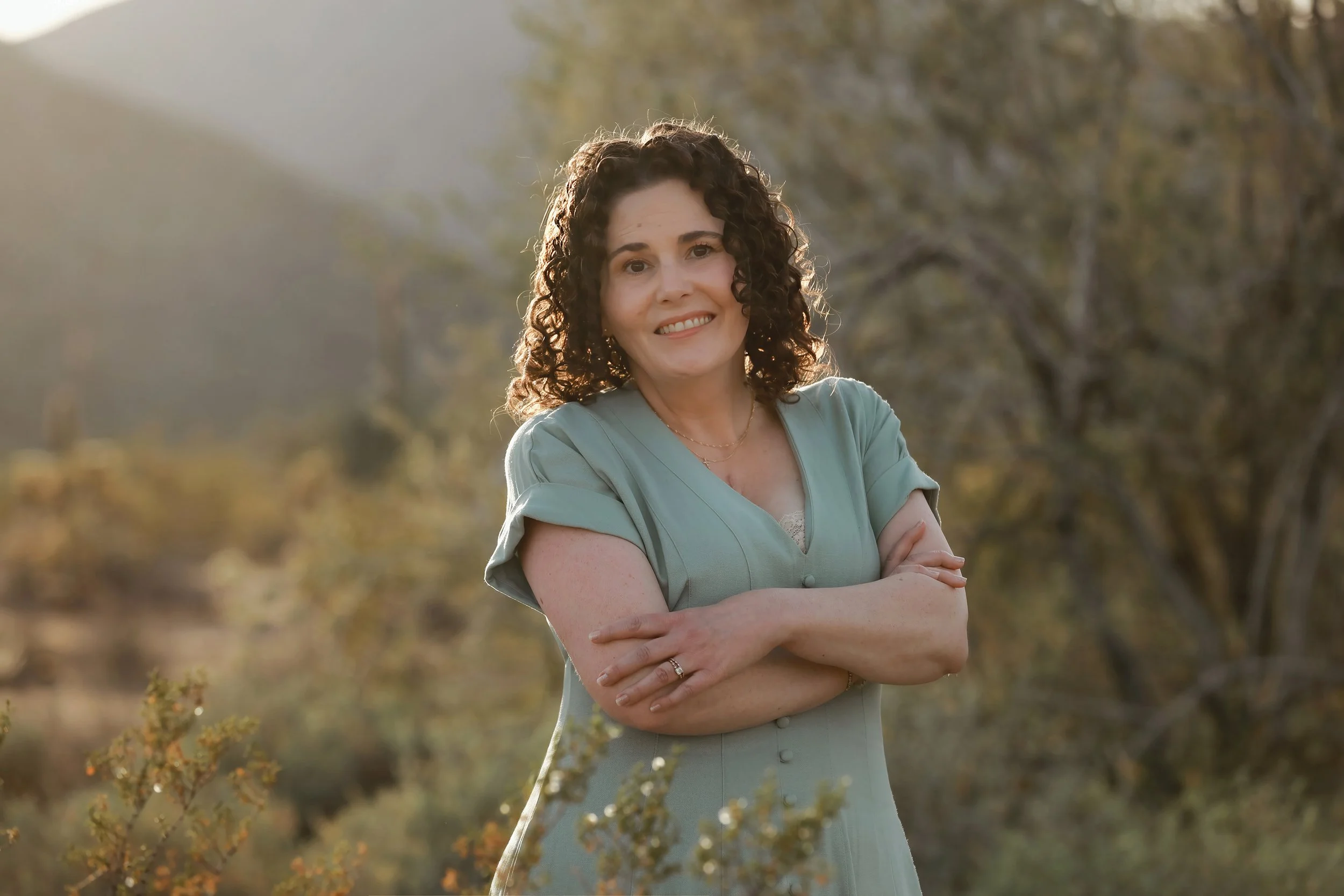 A woman with curly dark hair smiling outdoors with her arms crossed, in front of trees and foliage during daytime.