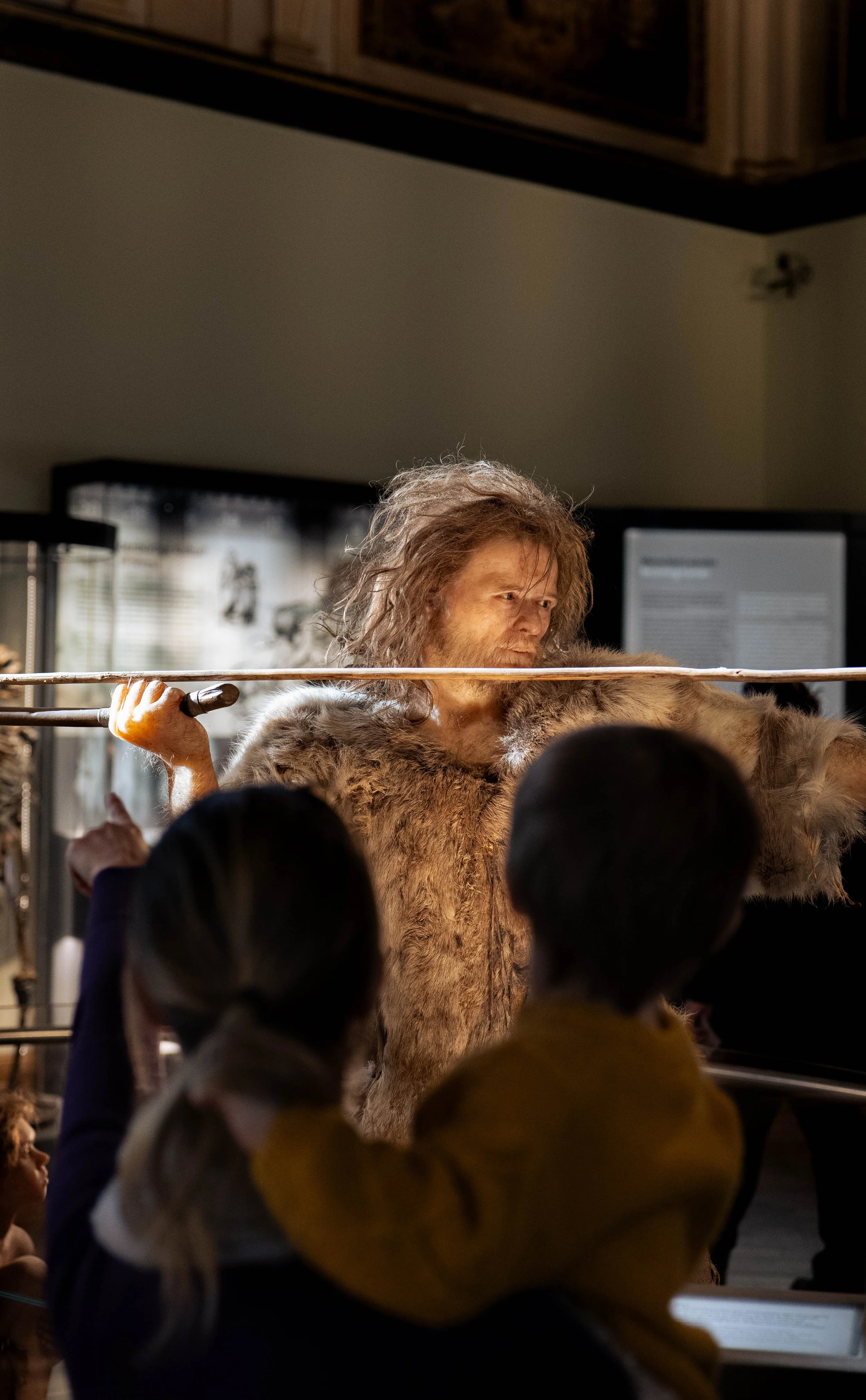 A person dressed as a prehistoric human in a fur outfit holding a spear and engaging with children at a museum exhibit.