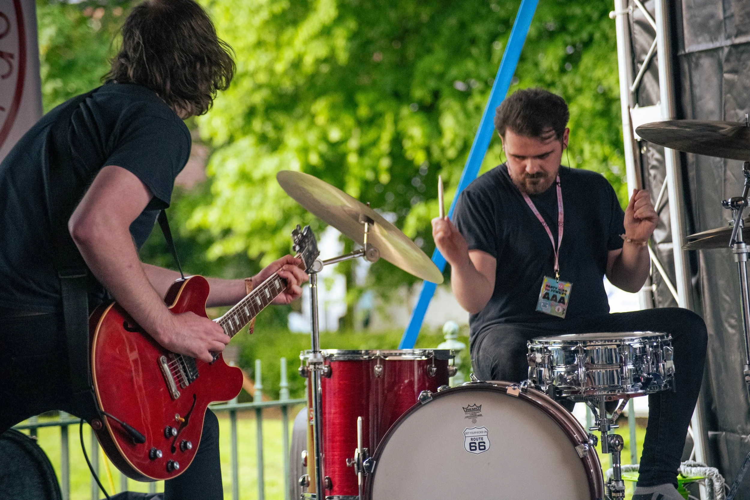 Two musicians playing instruments outdoors: one with a red electric guitar and the other on drums, in front of green trees.
