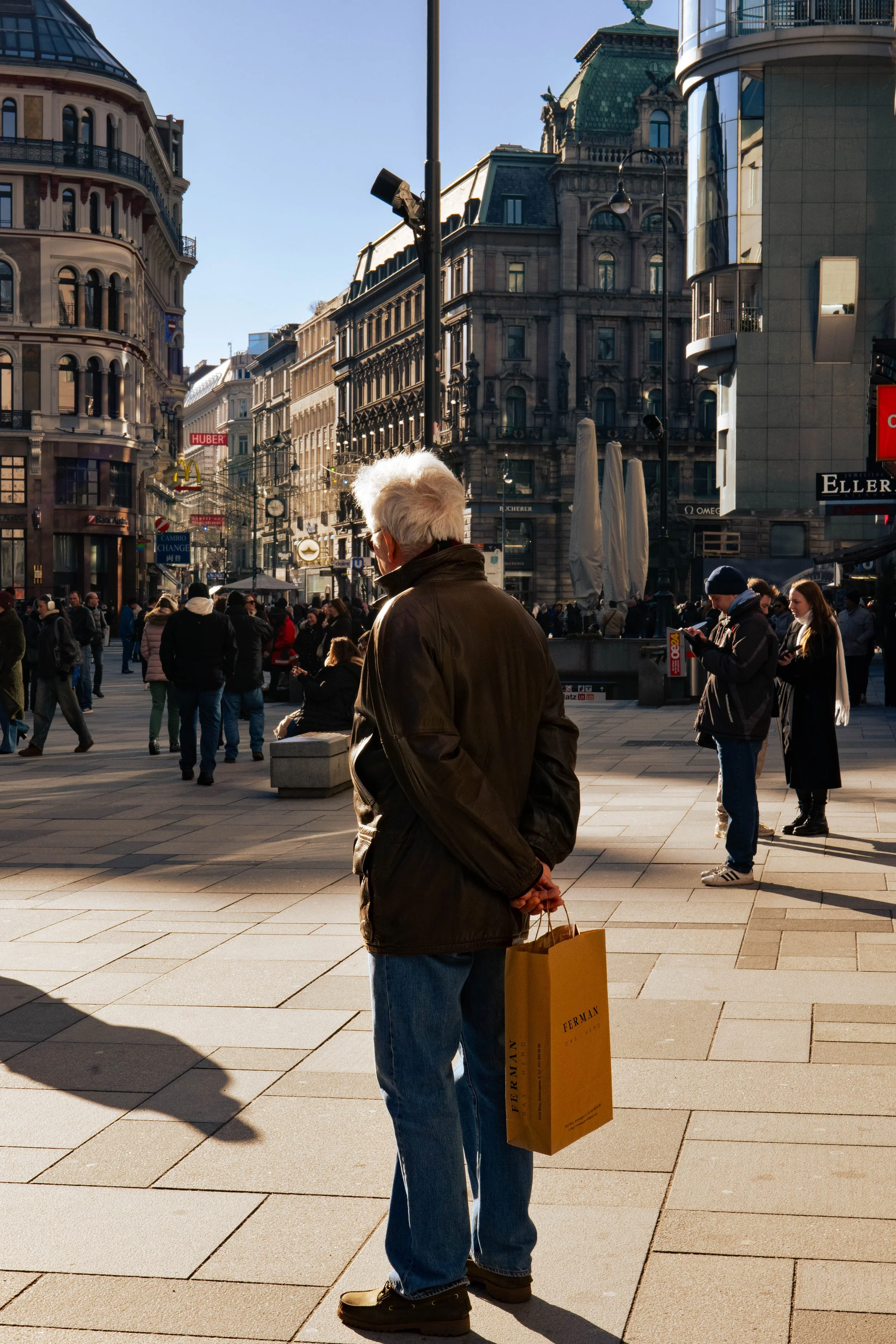 An elderly man with white hair, wearing a dark jacket and jeans, holding a yellow shopping bag, standing on a busy city street with many pedestrians and historic buildings in the background.
