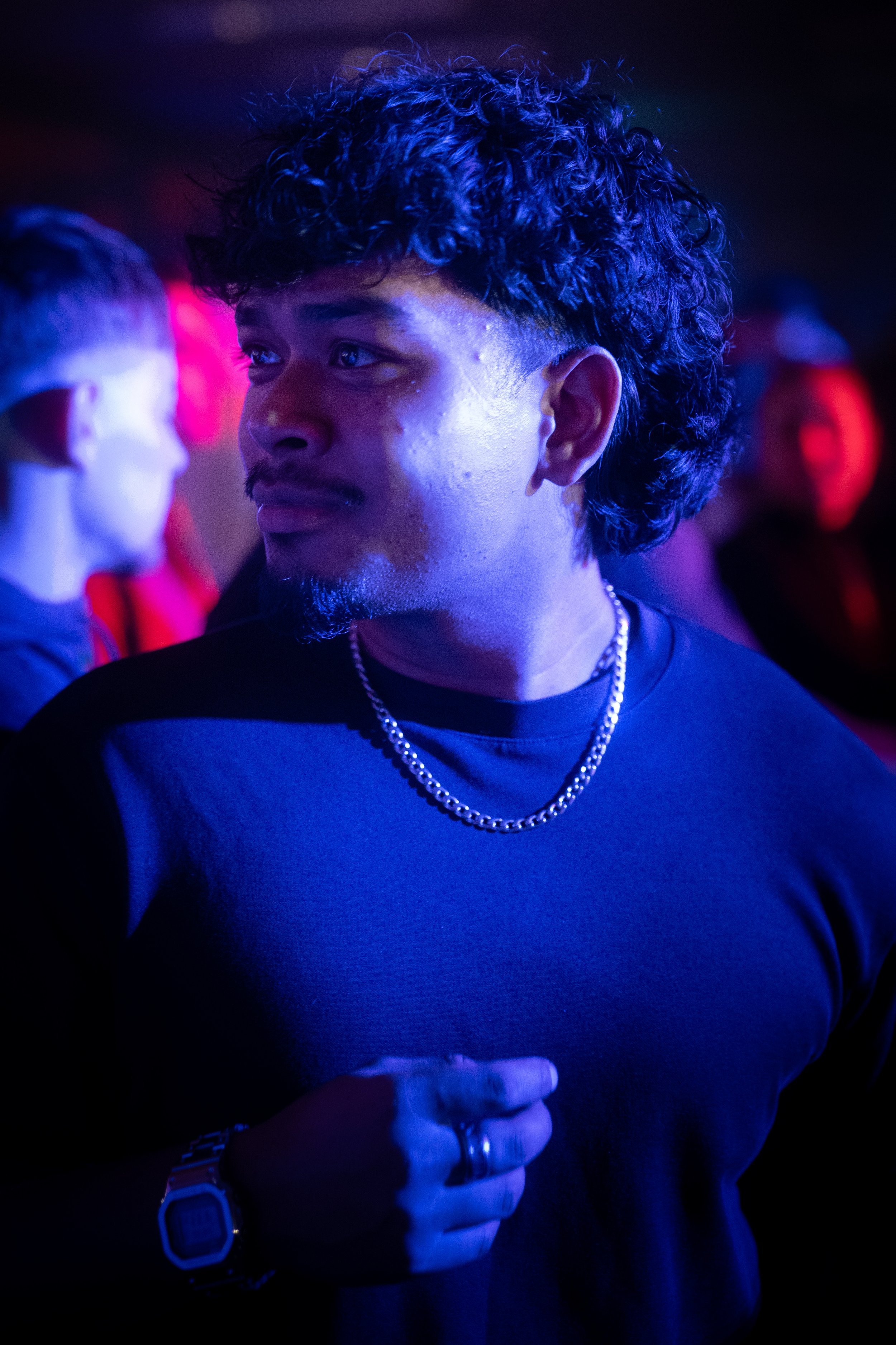 A man with curly dark hair, wearing a black shirt, a silver chain necklace, and a digital watch, standing in a dimly lit environment with blue and red lighting, surrounded by other people.