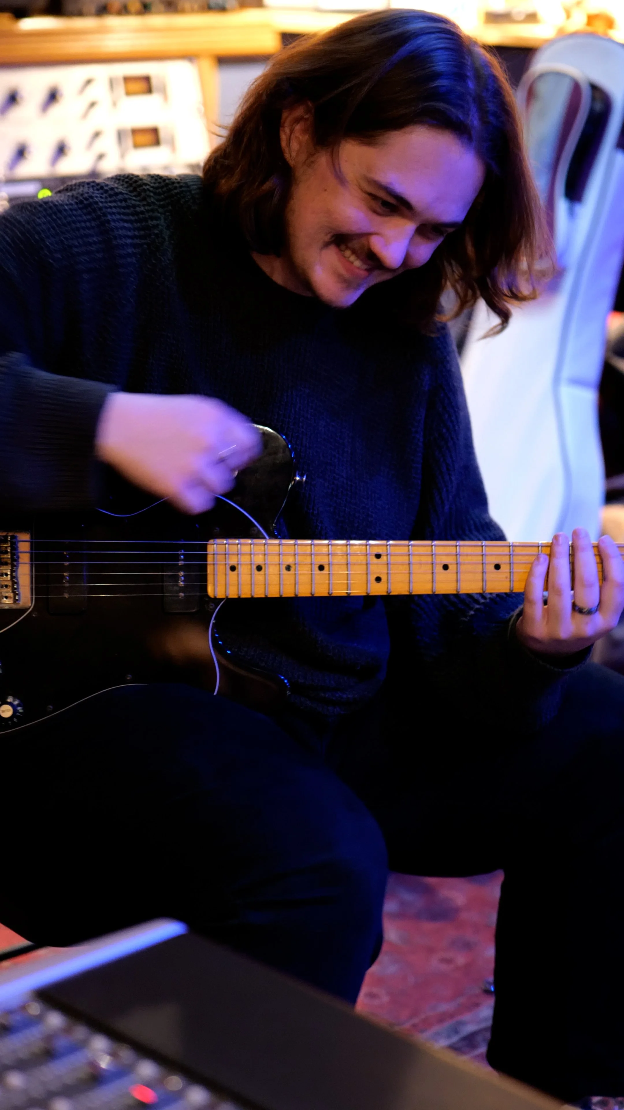 A young man smiling while playing a black electric guitar with a yellow fretboard, seated in a dimly lit room with colorful lighting.