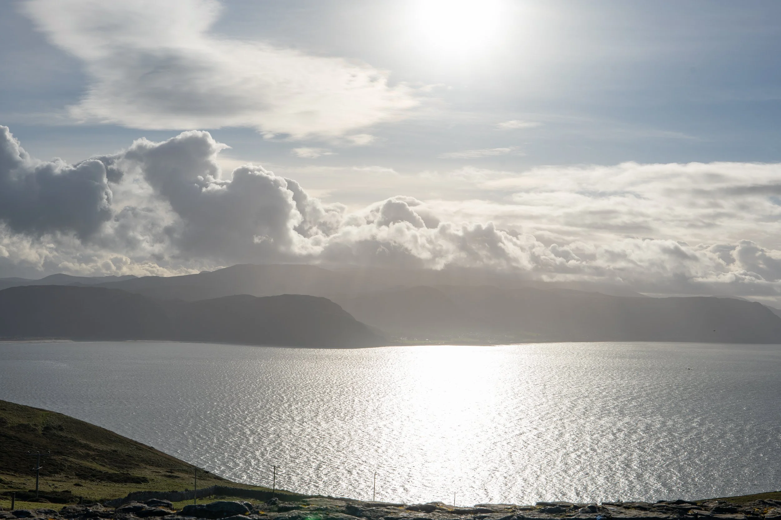 A scenic view of a large lake or sea with sunlight reflecting on the water, surrounded by mountains and cloudy skies.