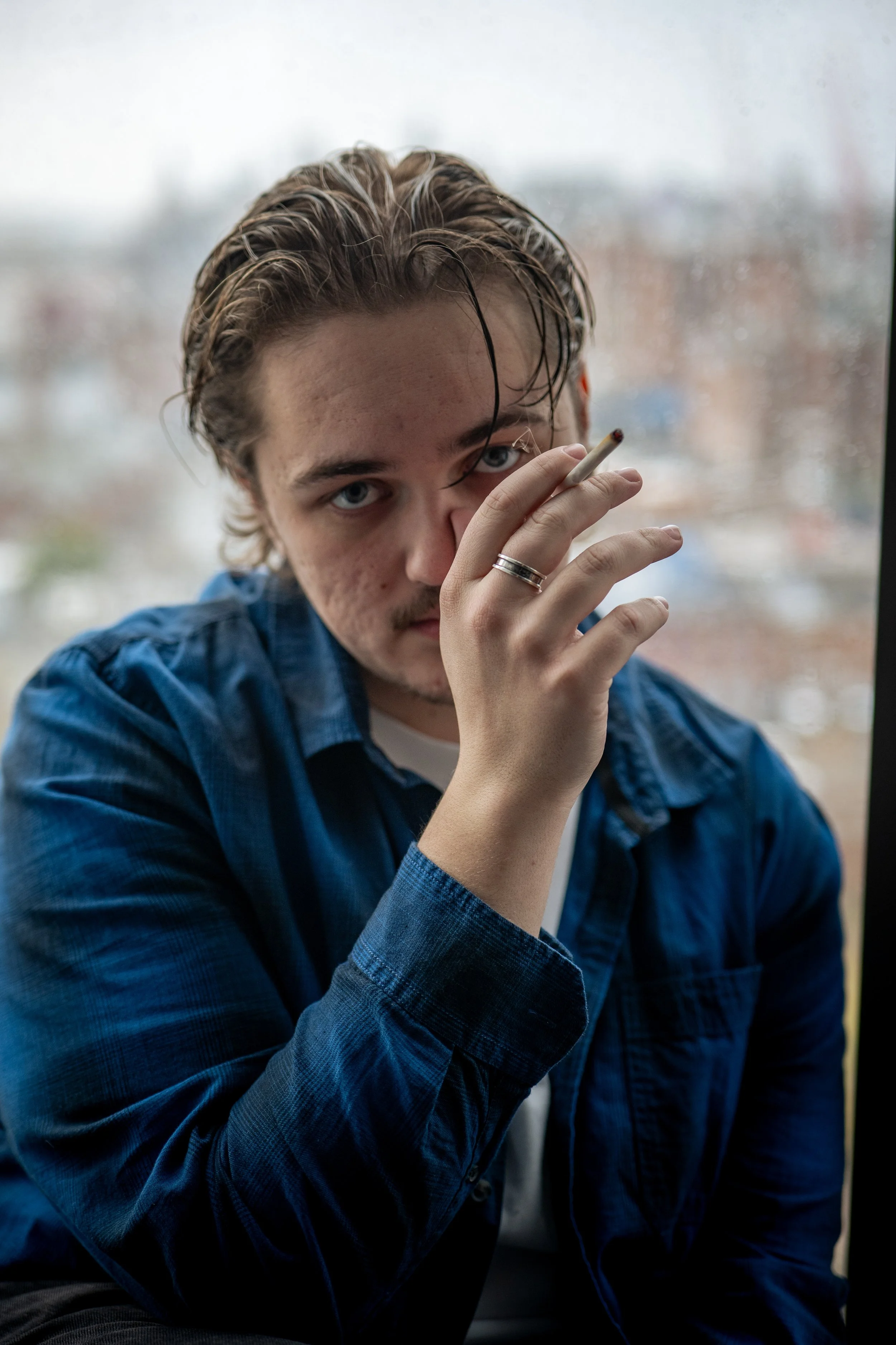 Young man with wet hair wearing a blue shirt, holding a cigarette near his face, looking directly at the camera, standing in front of a window with a blurry outdoor background.