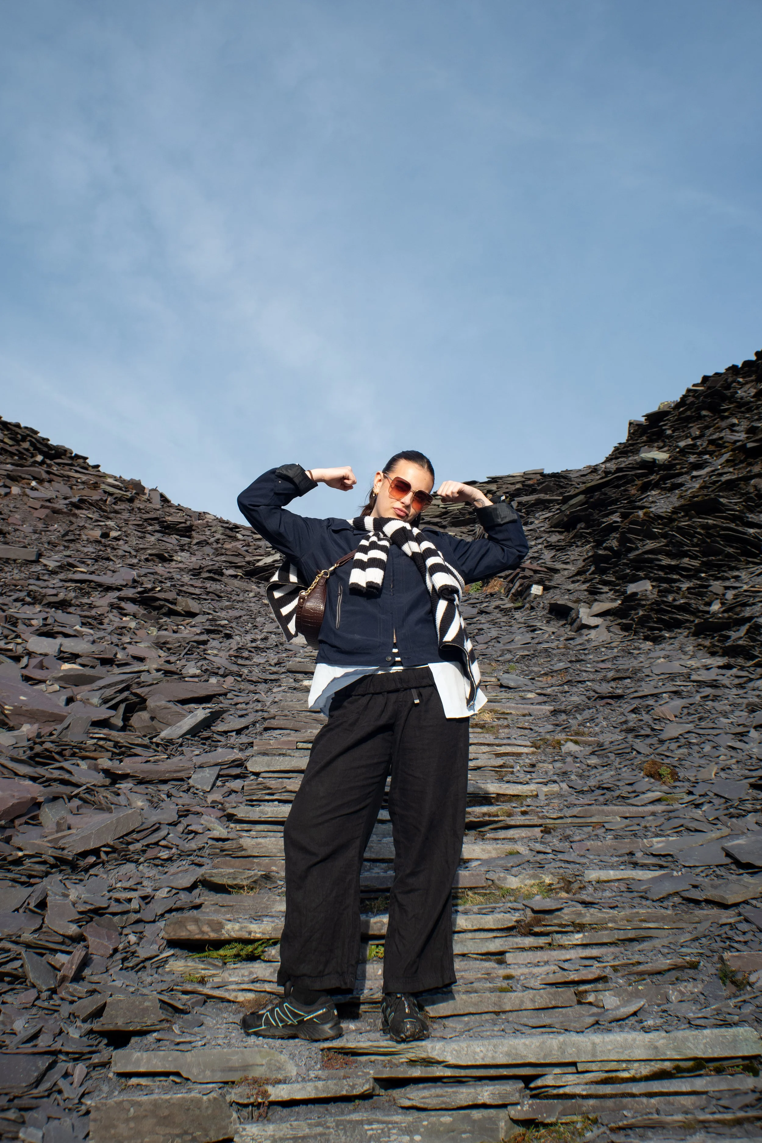 A woman standing on a rocky mountain trail, flexing her muscles, wearing sunglasses, a black jacket, a striped scarf, black pants, and hiking shoes.