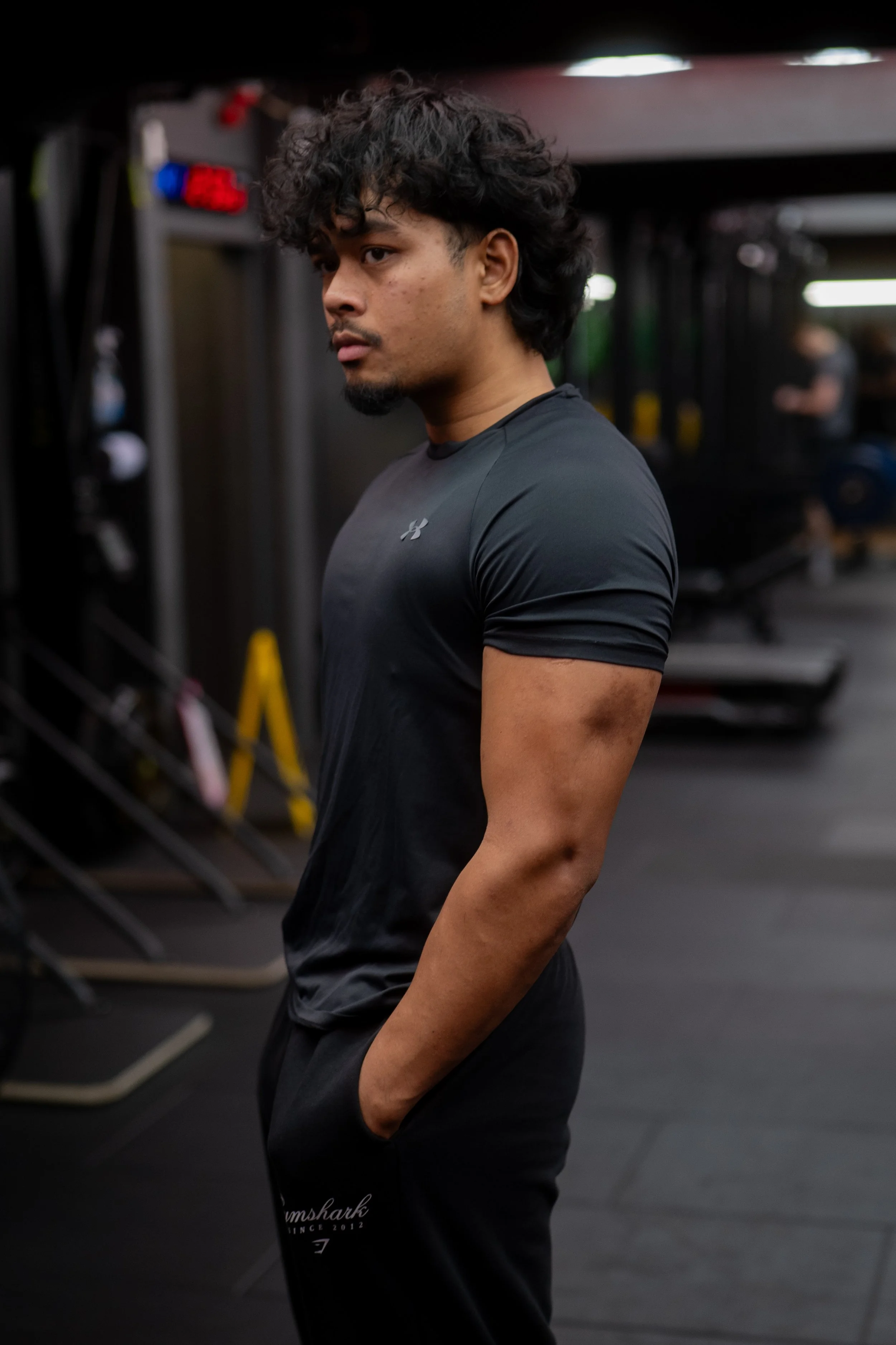 A young man with curly black hair and a goatee, wearing a black athletic t-shirt and black shorts, stands in a gym with his hands in his pockets, looking to the side.