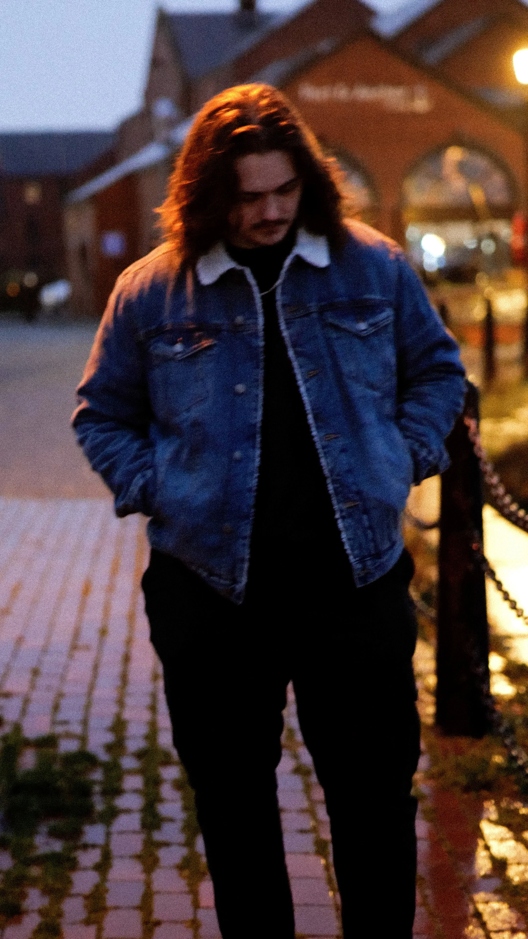 A man with long hair and a mustache wearing a denim jacket walking on a brick sidewalk at dusk in a small town.