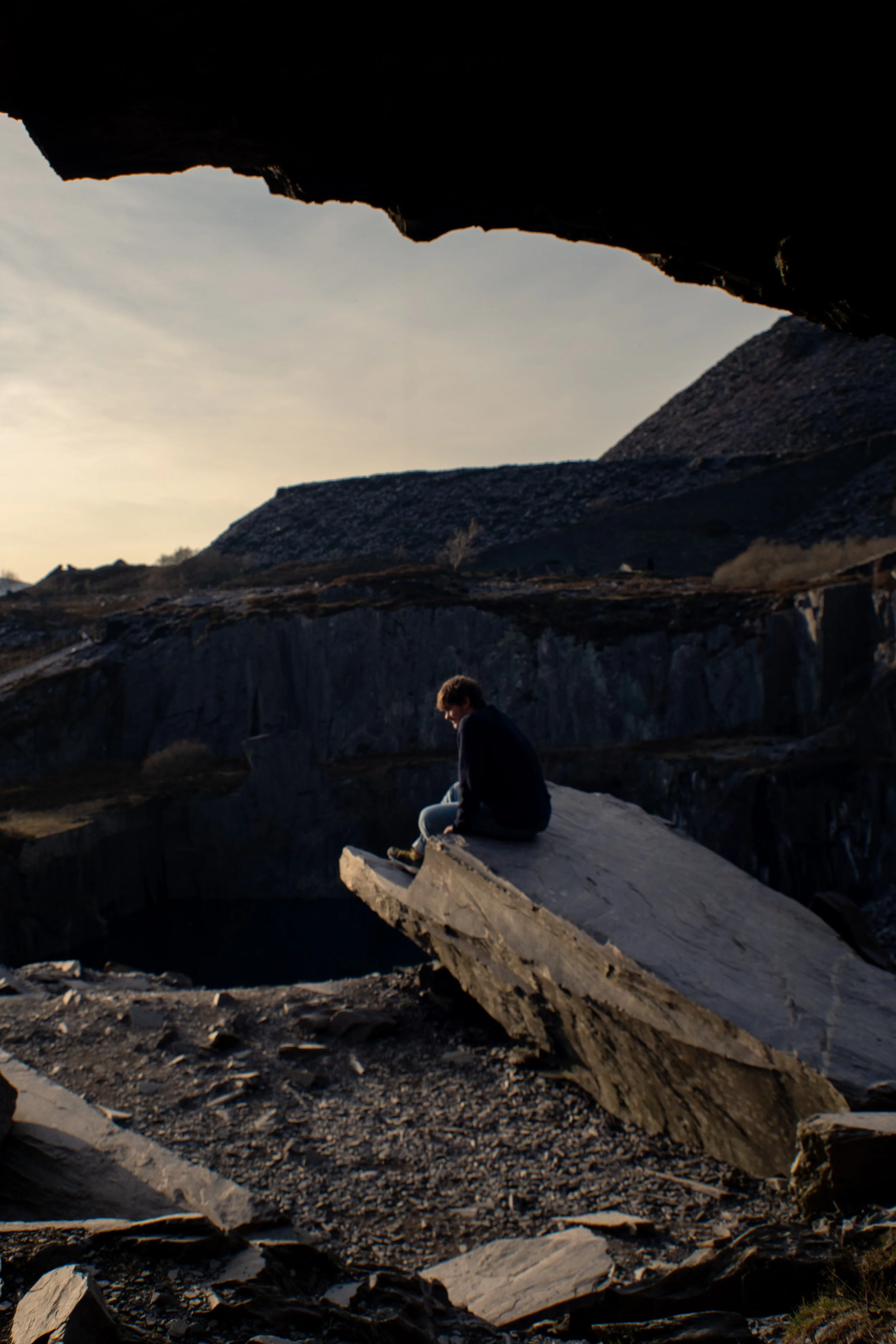 Person sitting on large rock formation in a rugged outdoor setting with rocky landscape and a canyon.