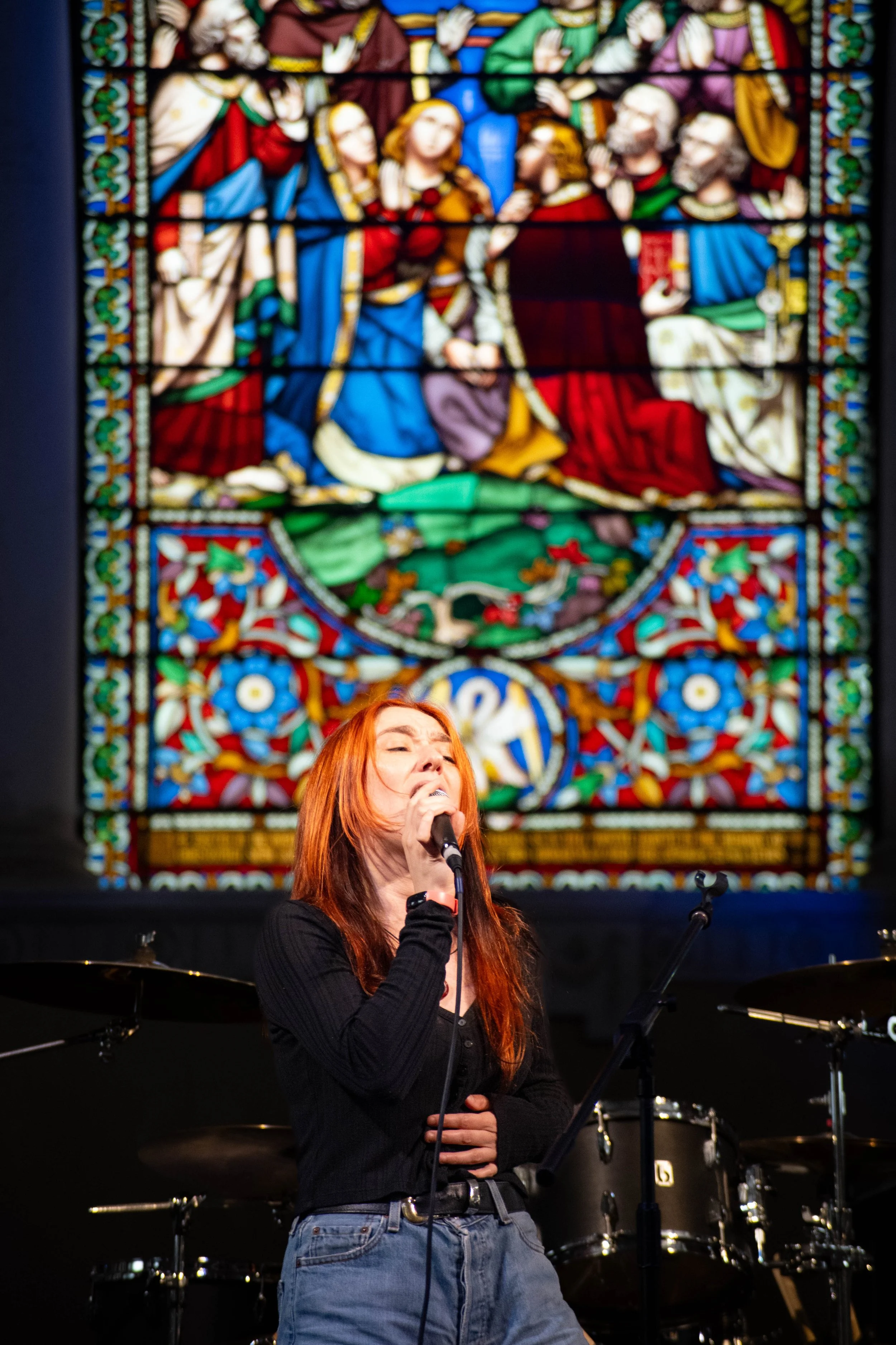 A woman with red hair singing into a microphone on stage, with a large stained glass window depicting a religious scene in the background.