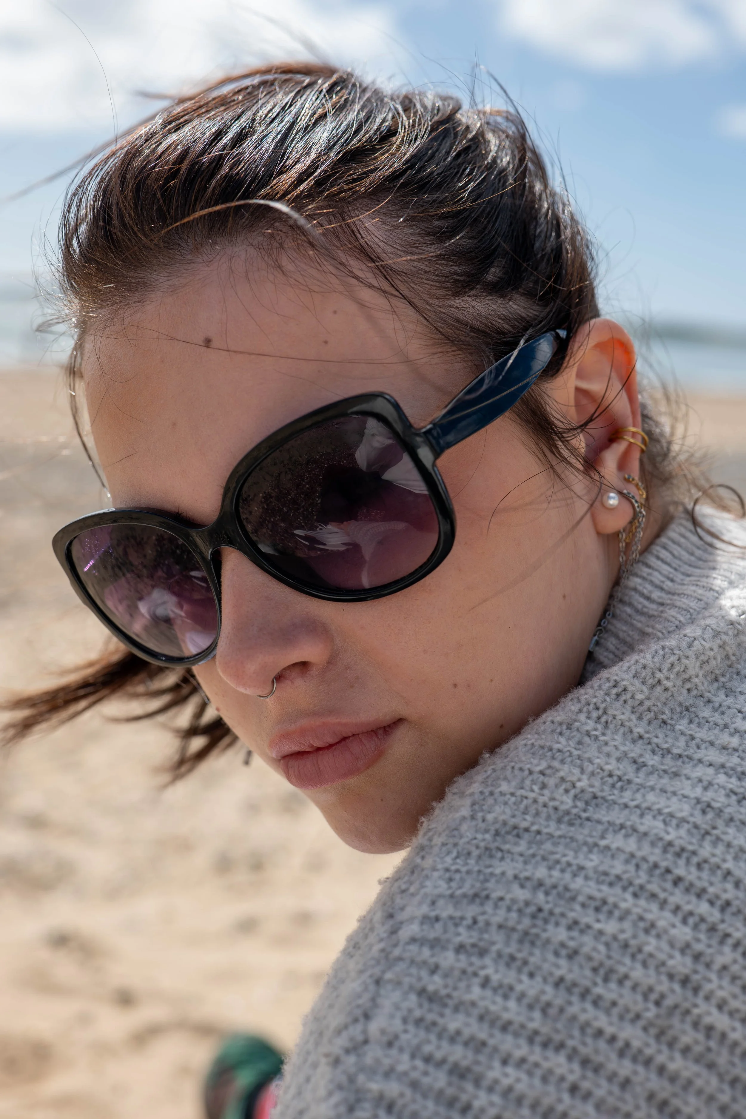 Close-up of a woman with short brown hair, wearing large black sunglasses, pearl earrings, a nose ring, and a gray knitted sweater, sitting on a sandy beach with water and blue sky in the background.