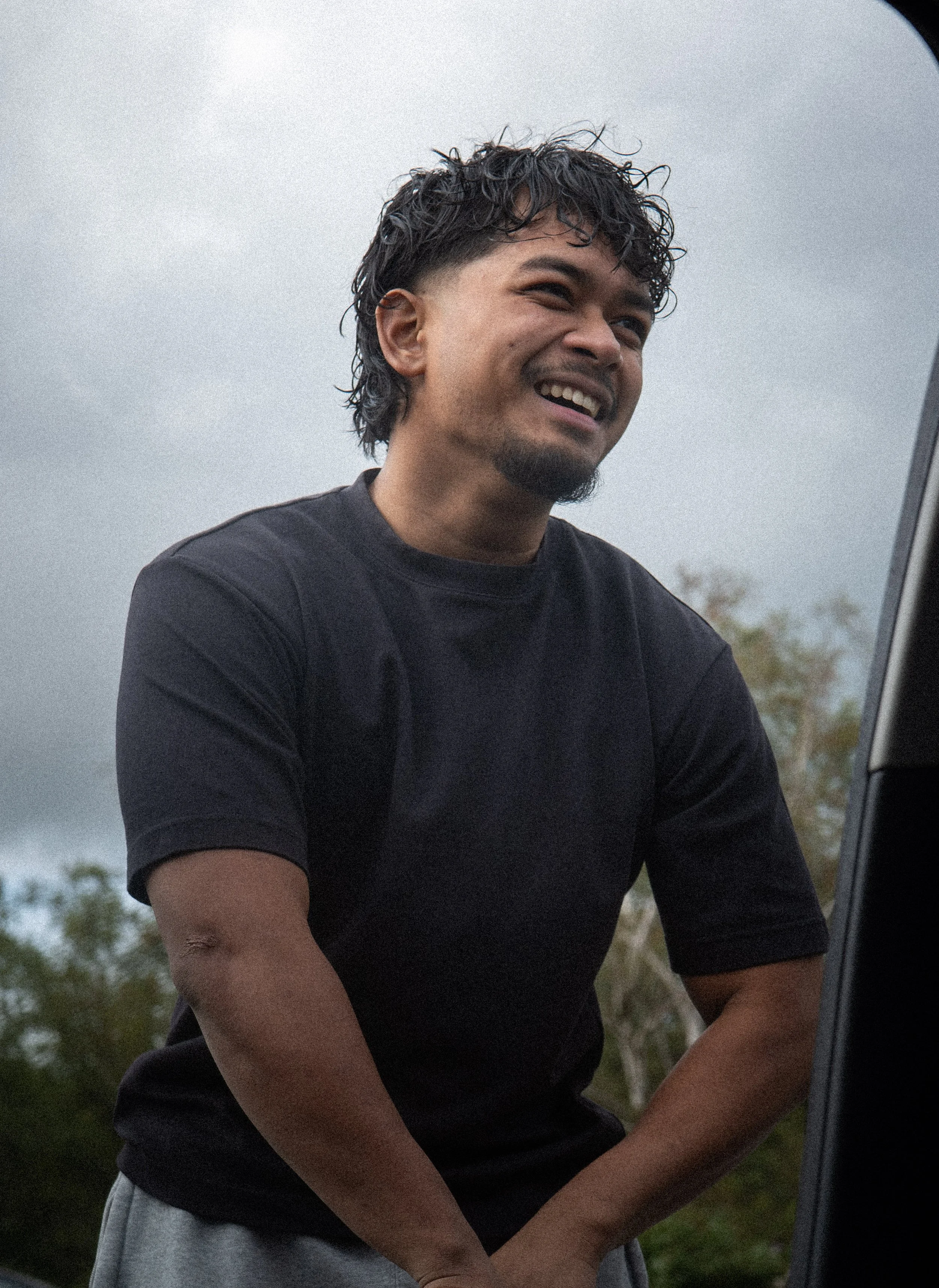 A man with curly black hair and a goatee, smiling, wearing a black T-shirt, standing outdoors near a car with a cloudy sky and trees in the background.