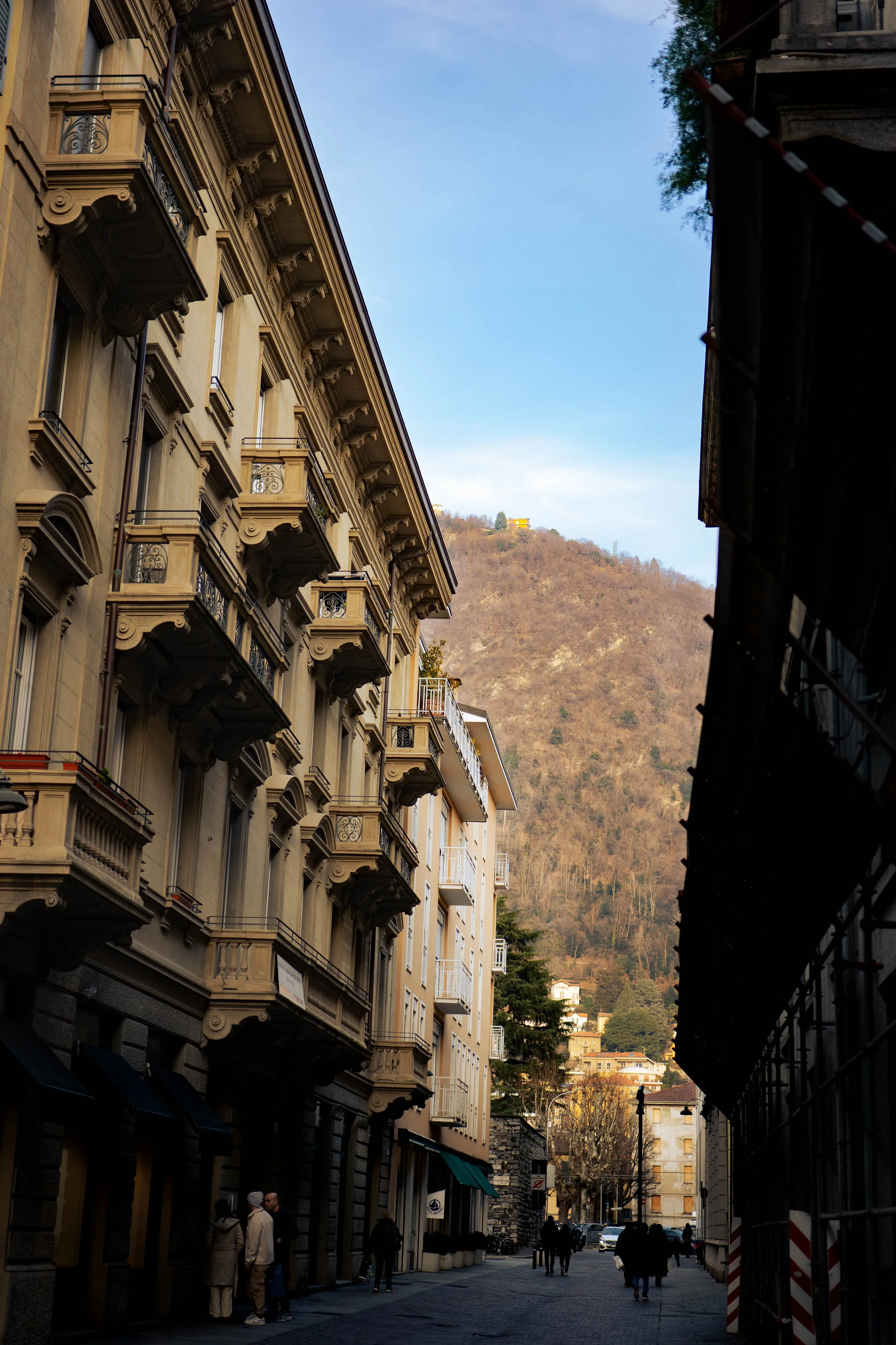 A street scene in a European city with tall, ornate buildings on the left, a mountain in the background, and a partly cloudy sky.