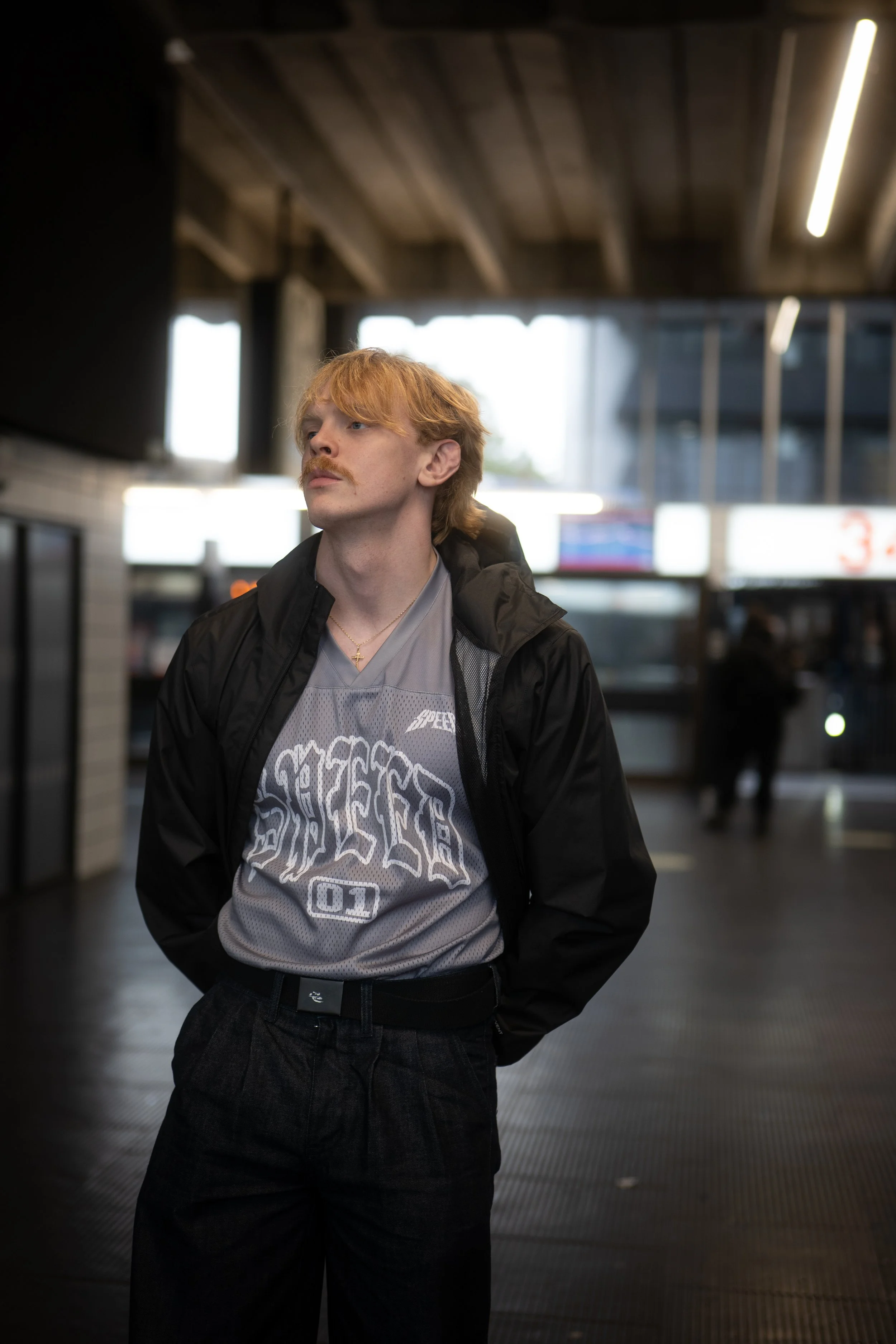 A young man with light skin, red hair, and a mustache, wearing a black jacket over a gray sports jersey, standing in an indoor public space, possibly a train station or airport, with his hands in his pockets, looking slightly upward to the left.