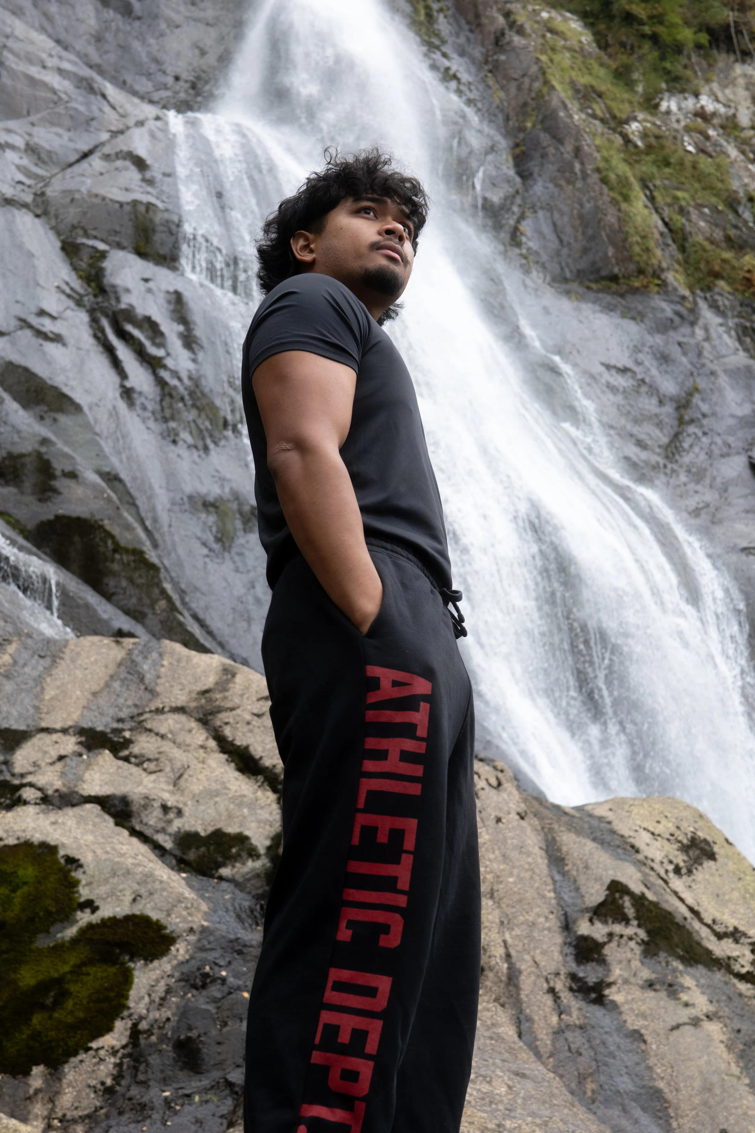 A man standing outdoors with a waterfall behind him, wearing a black t-shirt and black sweatpants with red lettering that reads 'ATHLETIC DEP'.