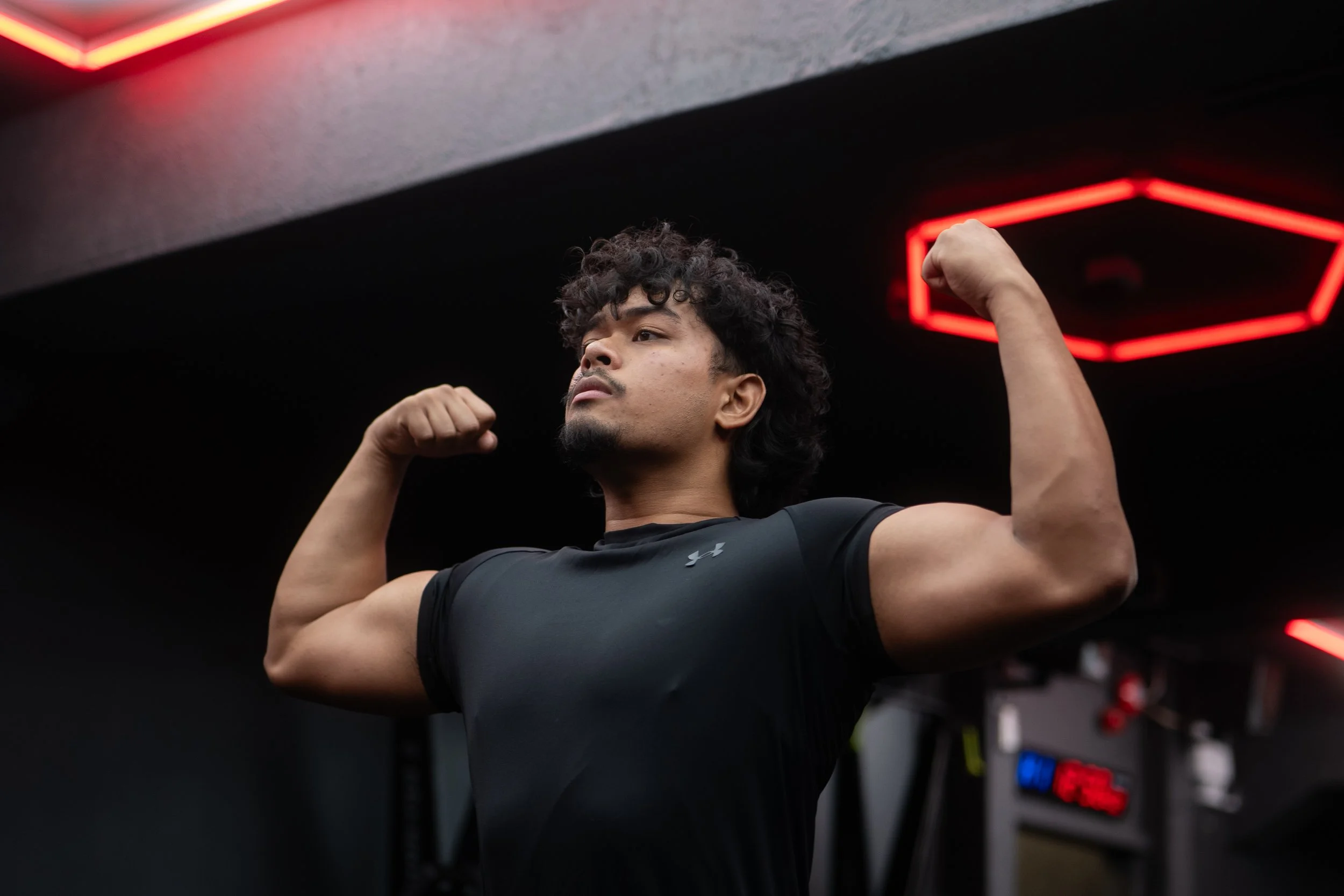 Young man flexing his biceps in a gym with red neon lights overhead.