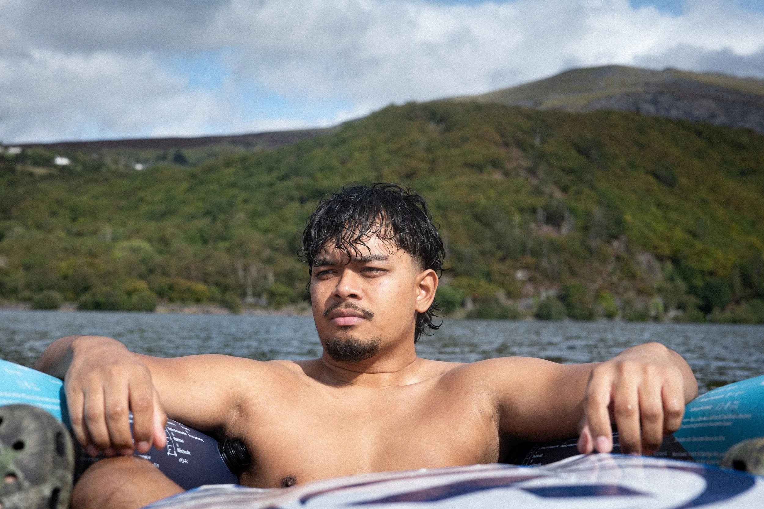 A shirtless man with wet curly hair relaxing on a float in a body of water, with green hills and cloudy sky in the background.