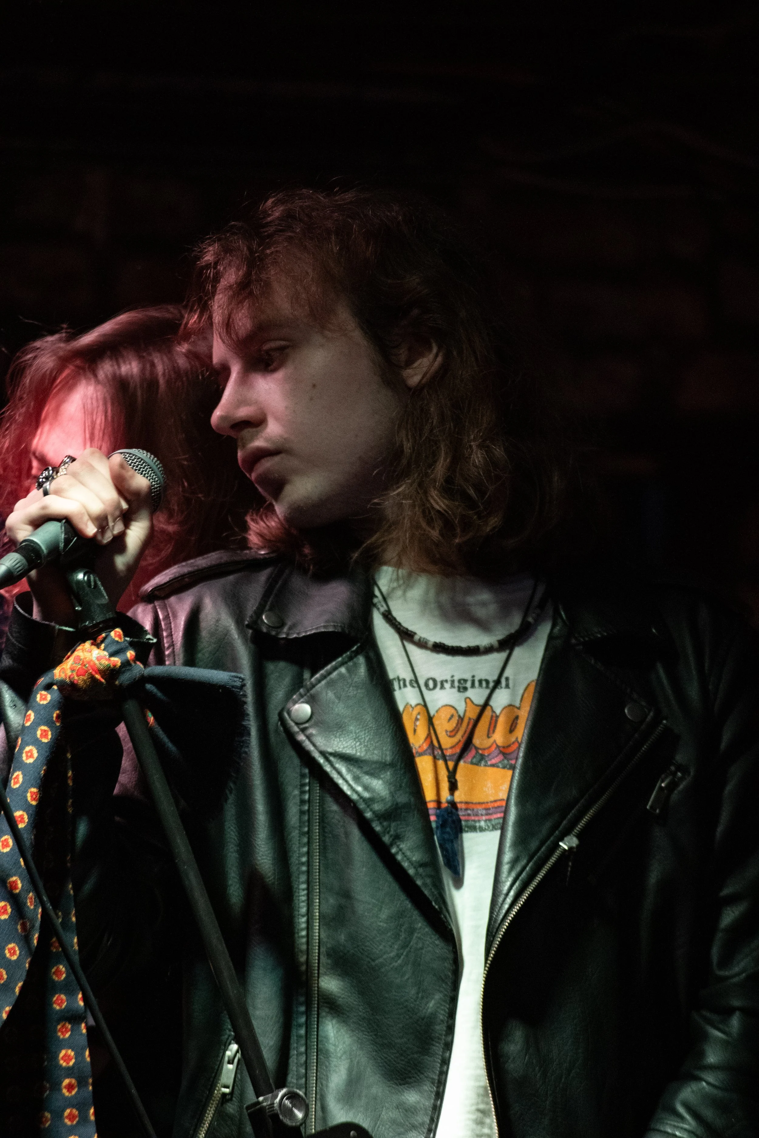 A young person with long, wavy hair holding a microphone on stage, wearing a black leather jacket and a graphic T-shirt, in a dark setting.