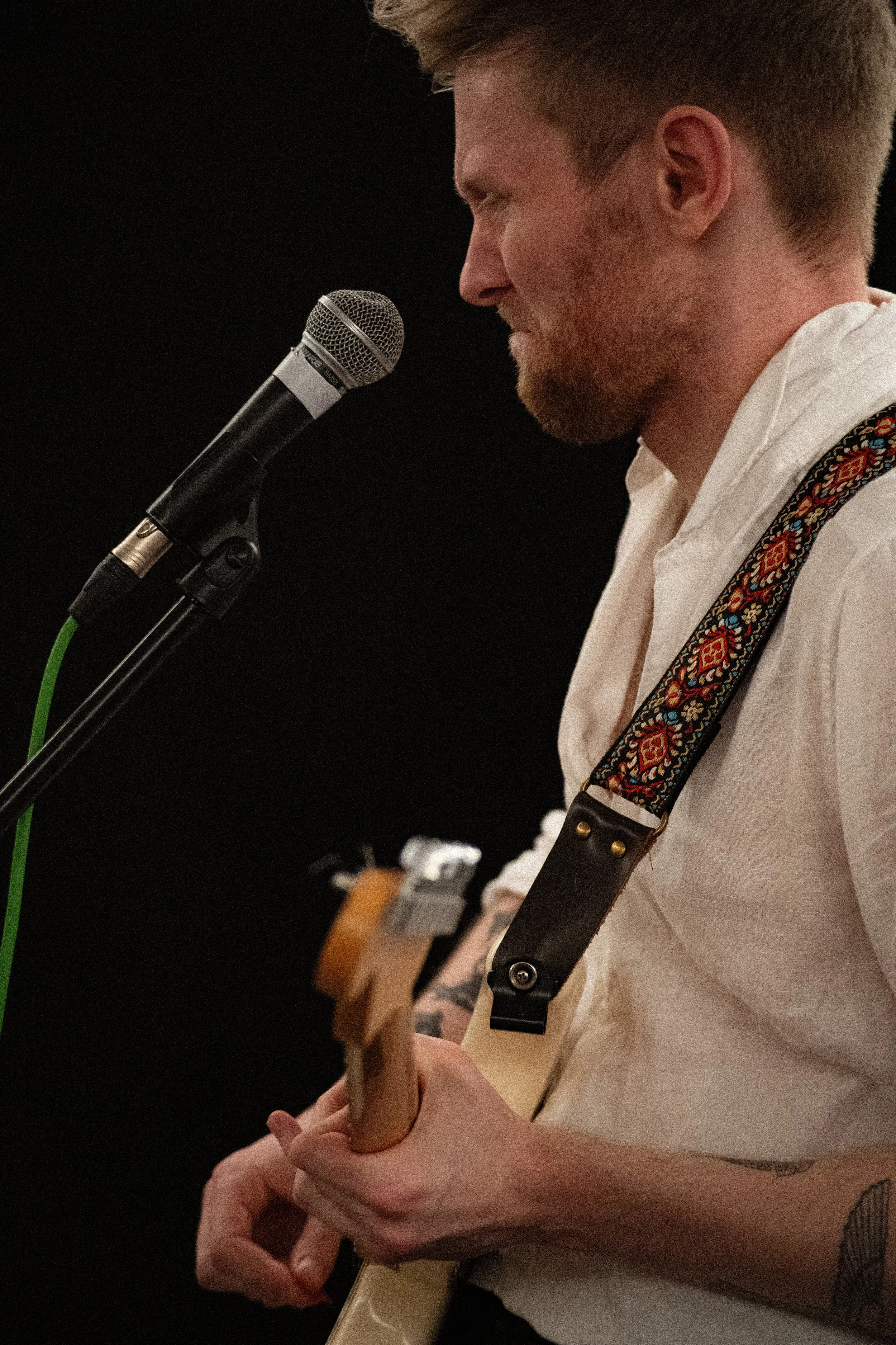 A young man with reddish-brown hair and a beard, wearing a white shirt with rolled-up sleeves, is playing an electric guitar and singing into a microphone against a black background.