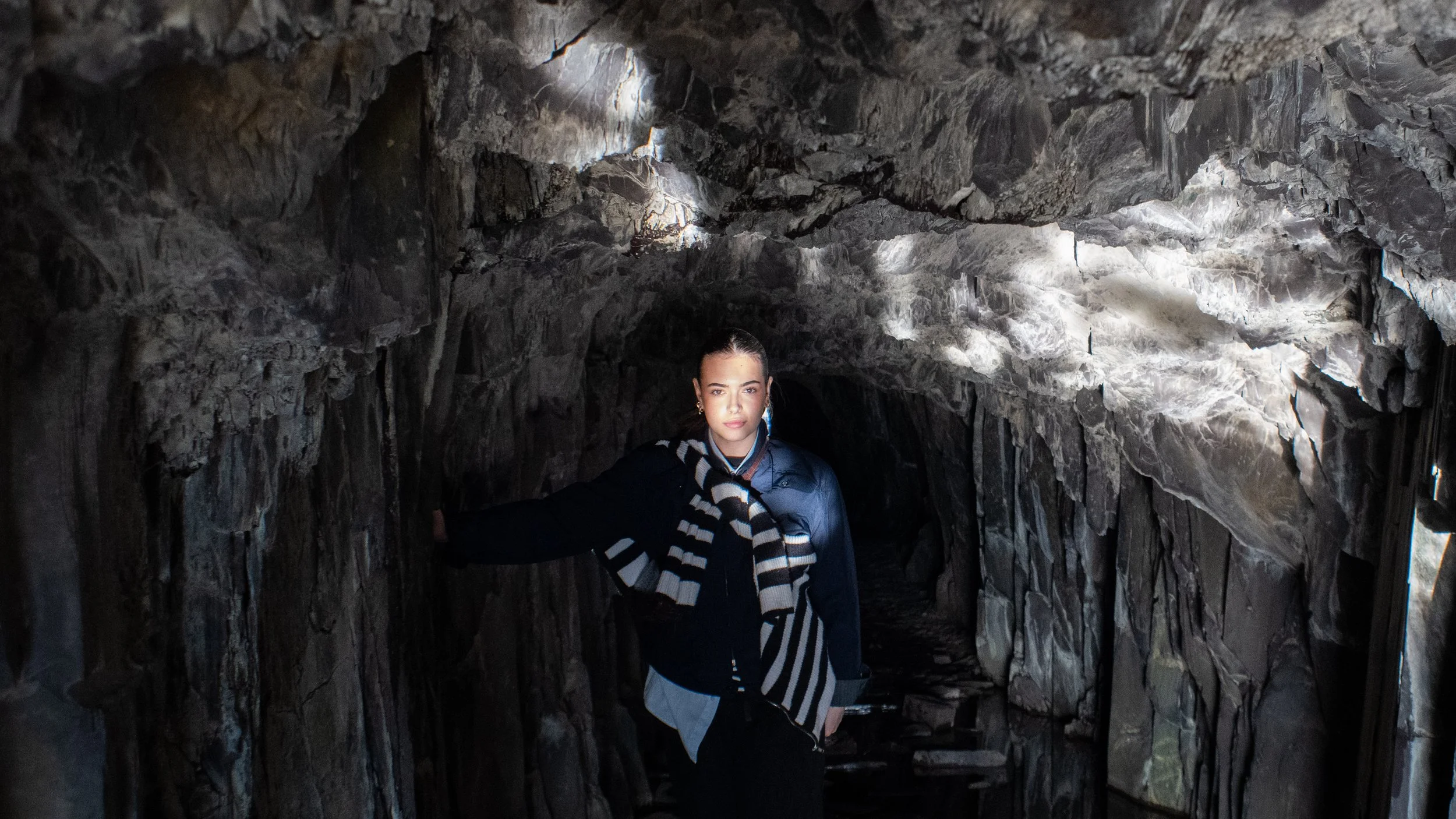 A young woman with dark hair pulled back, wearing a black jacket, striped scarf, and light blue shirt, standing inside a dark, rocky cave with uneven surfaces and some areas illuminated by natural light.