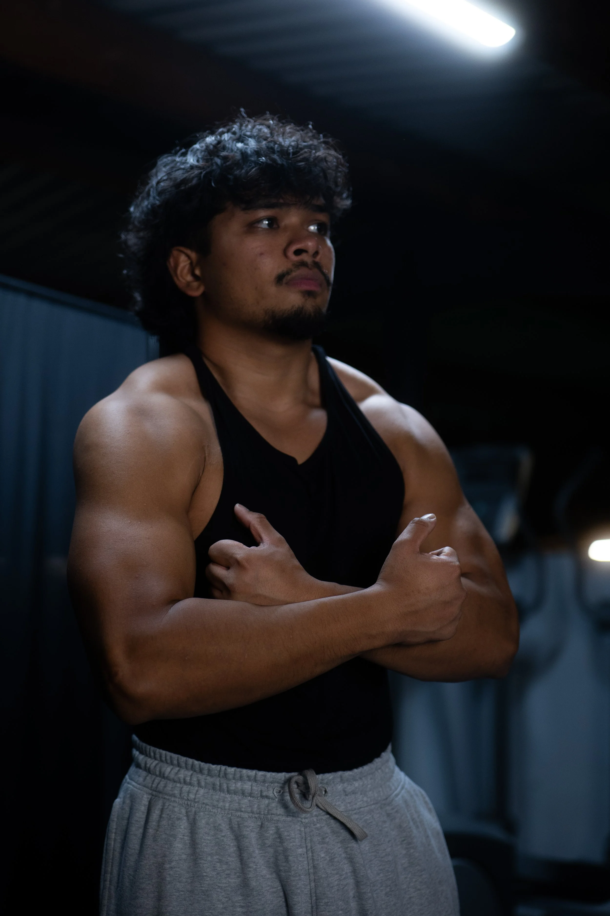 A muscular man with curly hair and a goatee standing in a gym, crossing his arms over his chest and looking off into the distance.