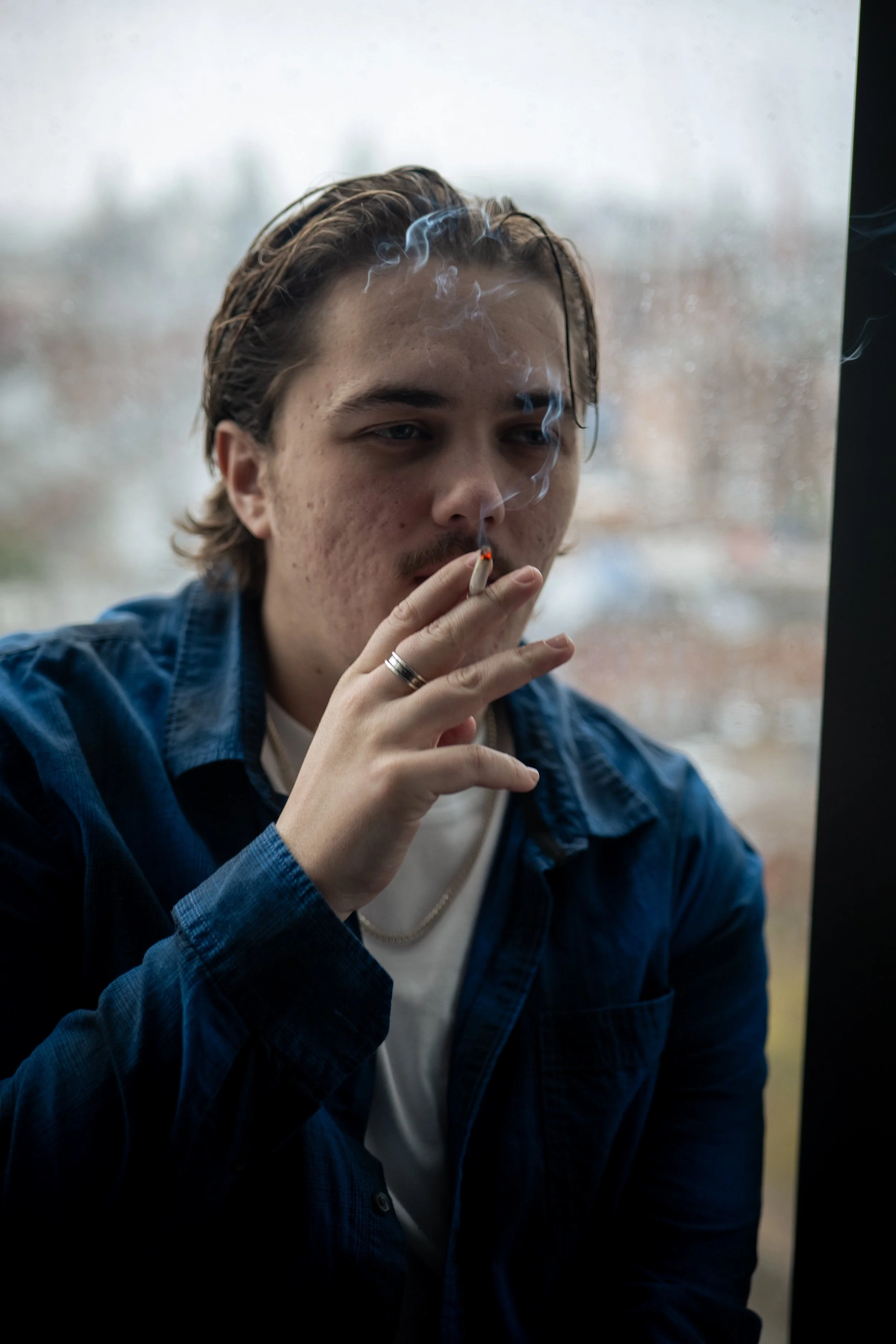 A young man with shoulder-length brown hair and a mustache smokes a cigarette near a window.