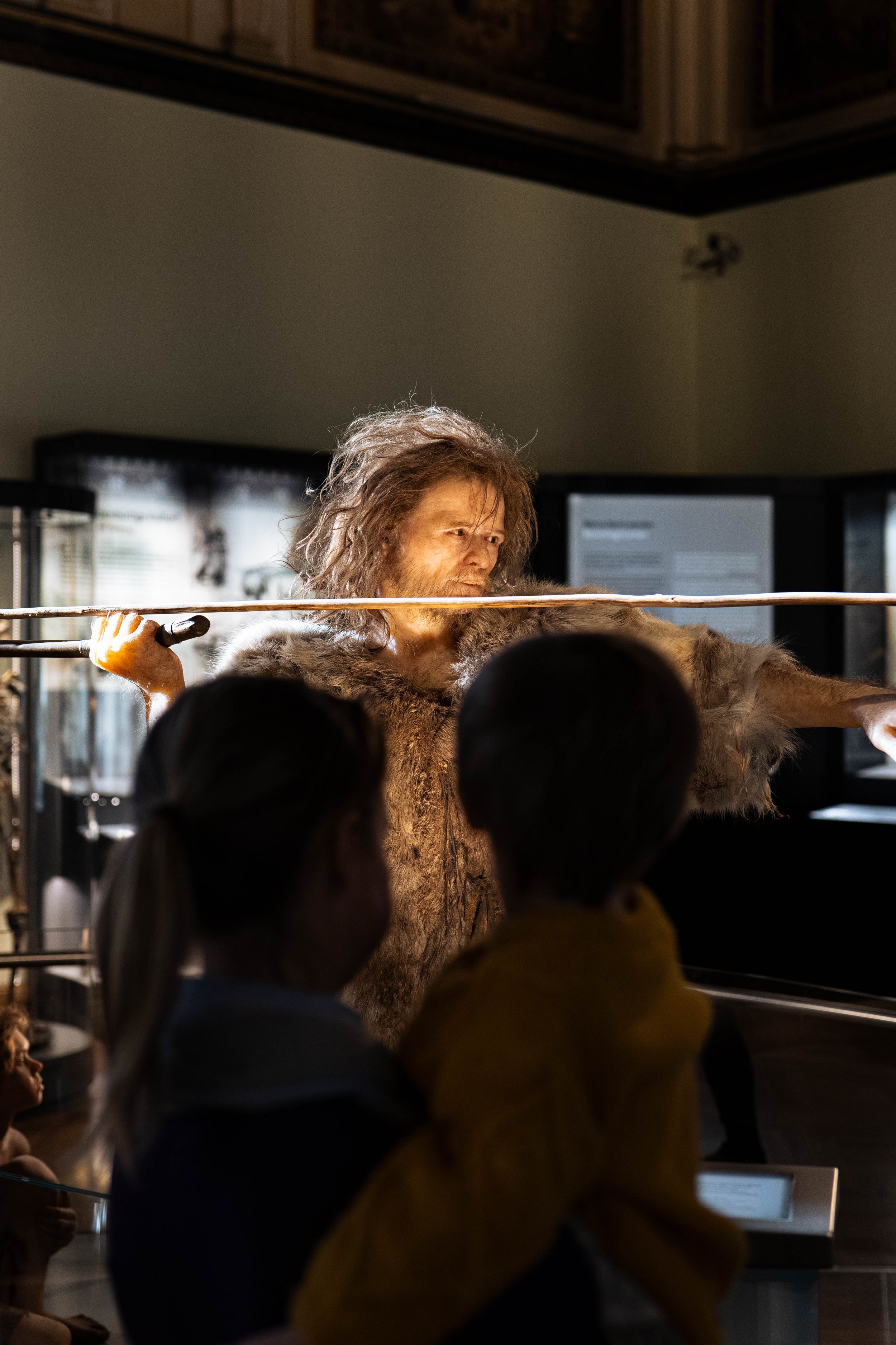 A museum exhibit features a person dressed as a prehistoric human, wearing animal fur and holding a spear, while two children watch.