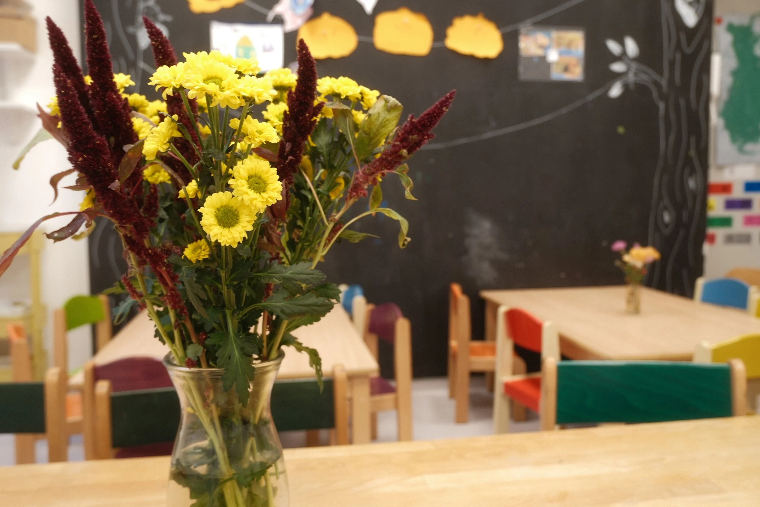 Foto von Wohnküche. Im Vordergrund ein Blumenstrauß mit gelben und roten Blumen in einer Vase, in einem Raum mit bunten Stühlen und einer schwarzen Wandtafel im Hintergrund.