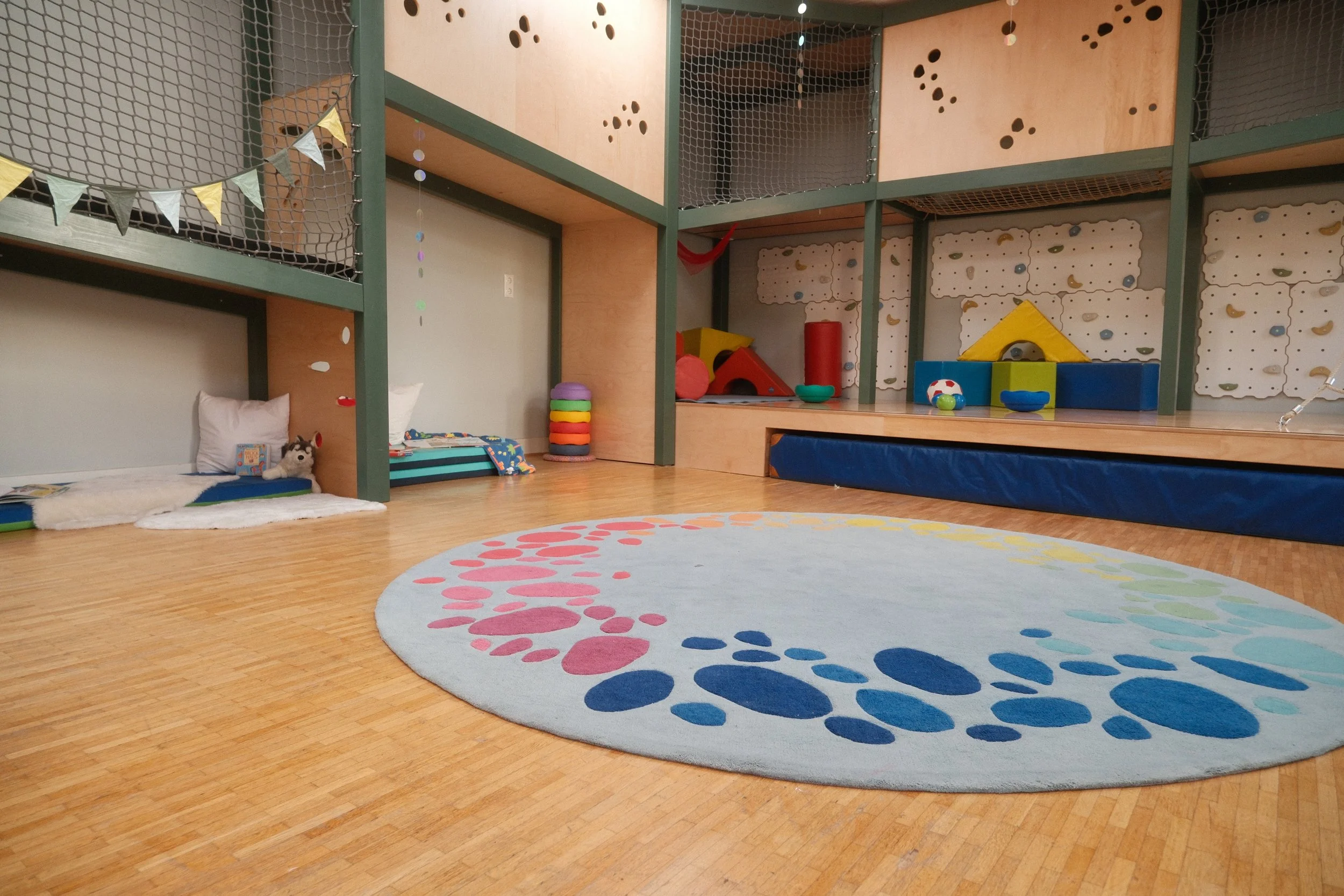 Foto Bewegungsraum, ein farbenfroher Spielraum für Kinder mit einem großen, runden Teppich in Pastellfarben, Klettermöglichkeiten, Spielzeug und Stapelsteinen.