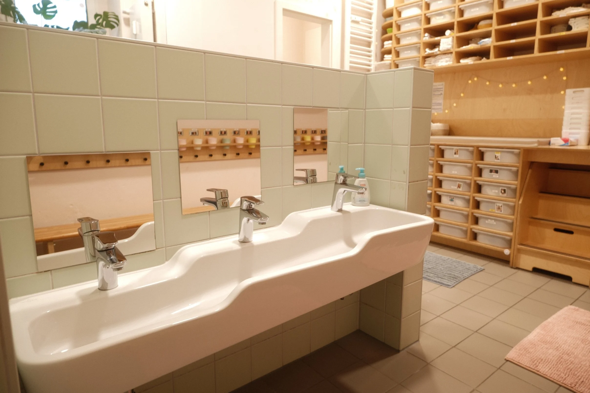 Foto vom Badezimmer, mit Waschbecken und Spiegeln auf unterschiedlichen Höhen. Im Hintergrund befindet sich eine Wickelkommode mit Aufbewahrungsboxen.