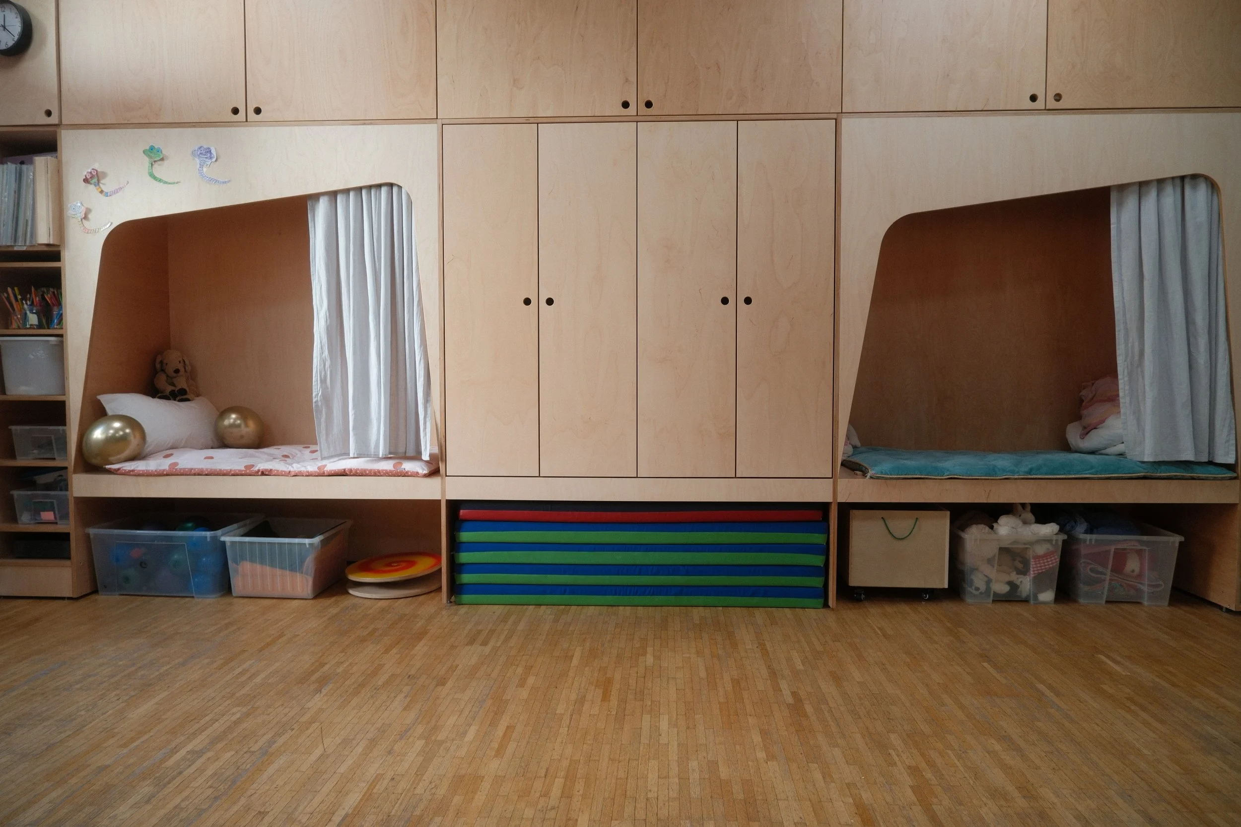 Foto des Bewegungsraums mit zwei Schlafnischen, jeweils mit Kissen, Vorhängen und Spielsachen, umgeben von Bücherregalen und Aufbewahrungsboxen, alles in einer individuell angefertigten Holzkonstruktion